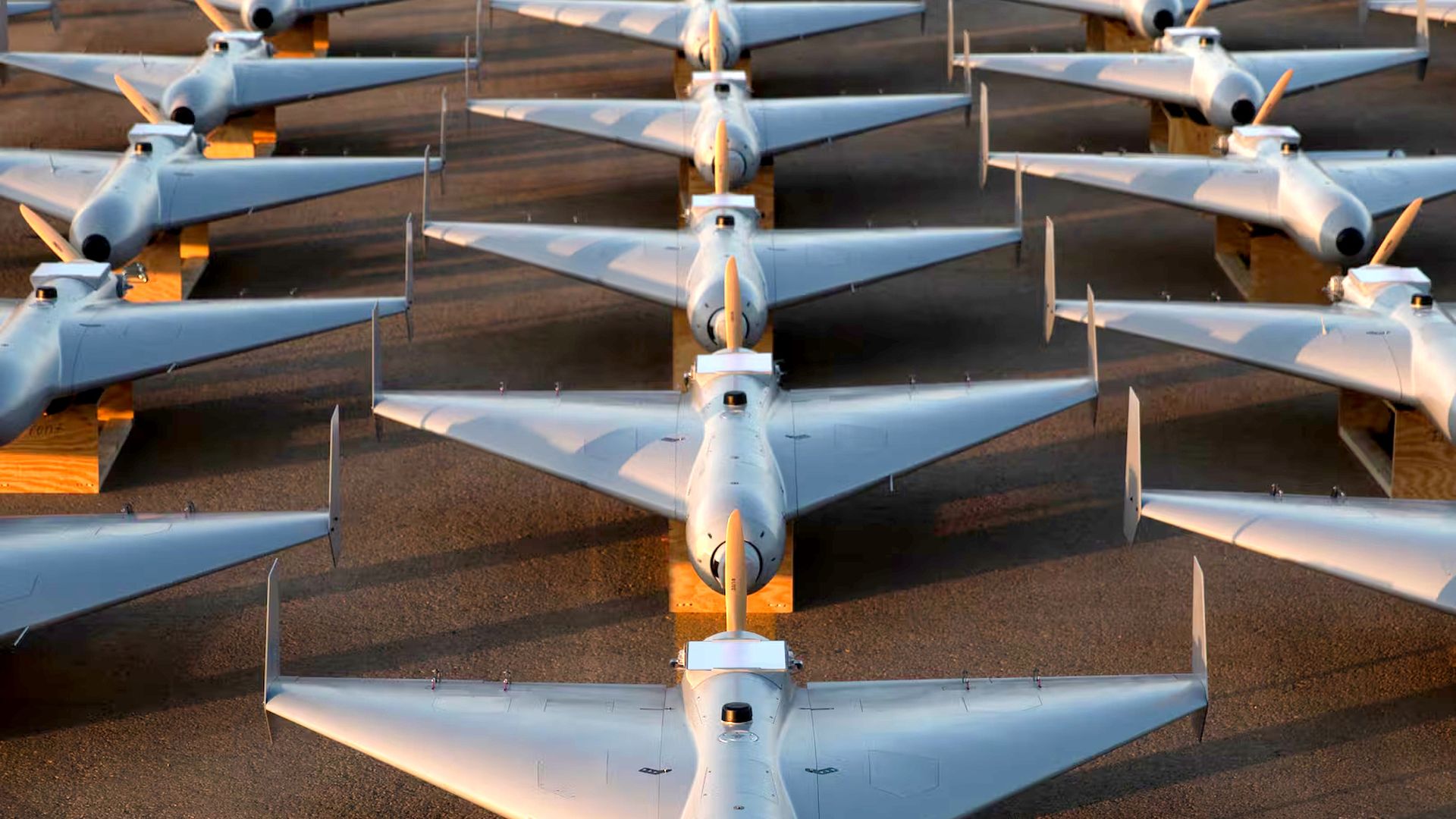 Rows of identical silver military drones with orange propellers aligned on the ground, bathed in warm sunlight, forming a symmetrical pattern.