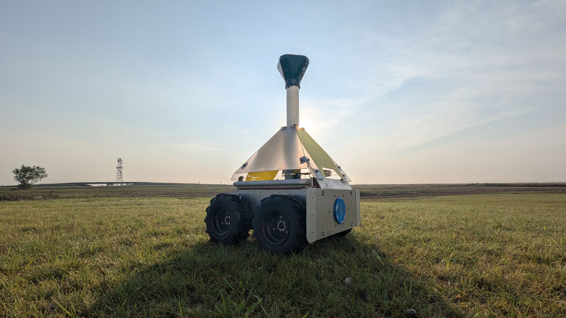 Four-wheeled autonomous rover with a tall black camera mast and white body sits in a grassy field under a clear blue sky; sun behind it, with a distant tower and a lone tree on the horizon.