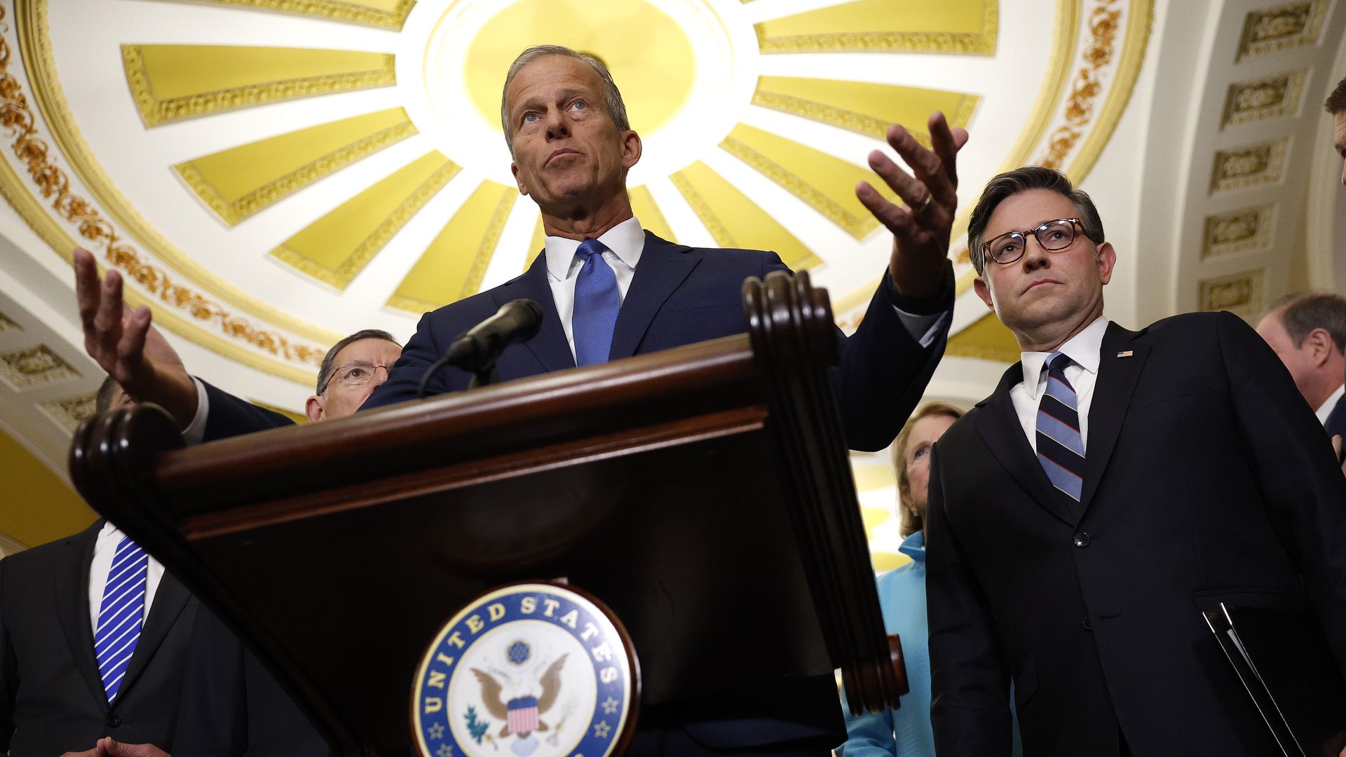 A man in a dark suit and blue tie speaks at a wooden podium bearing the U.S. Senate seal, flanked by colleagues in a gilded room with a gold sunburst ceiling.