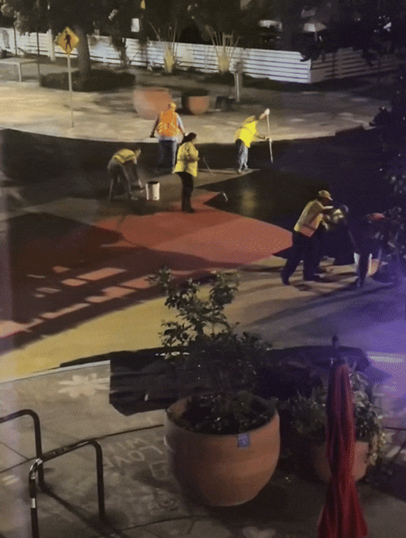 Workers in yellow reflective vests paint black over a rainbow-colored intersection.