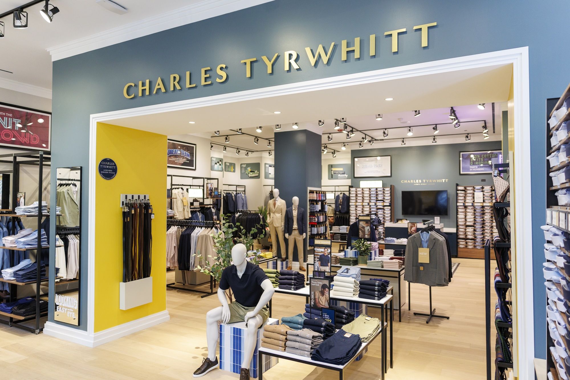 Interior of a Charles Tyrwhitt clothing store with blue walls, a yellow pillar, racks of suits and shirts, and mannequins near front tables under bright track lighting.