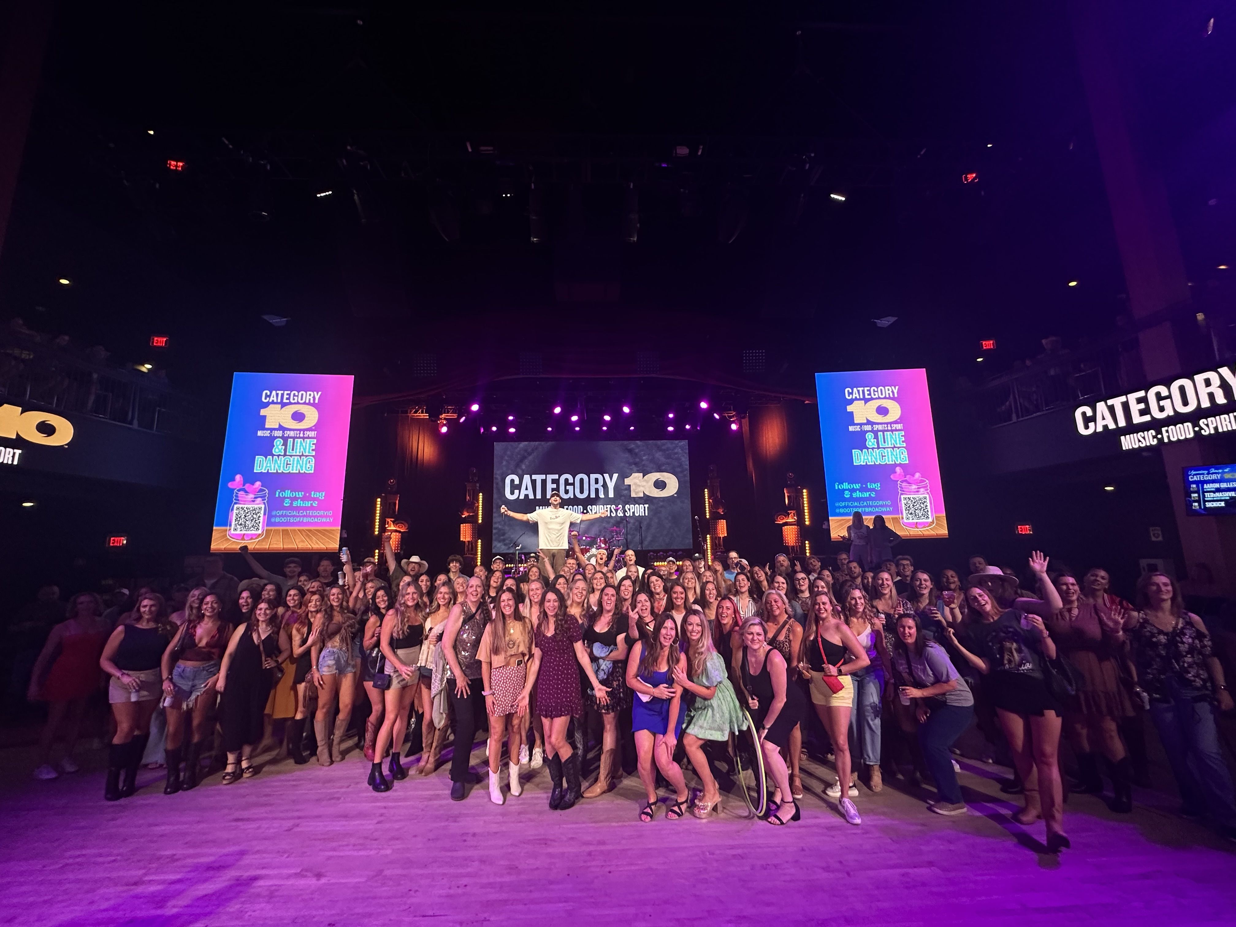 Large group of people posing and smiling in a dimly lit event venue with purple stage lights and screens displaying "CATEGORY 10 MUSIC-FOOD-SPIRITS & LINE DANCING."