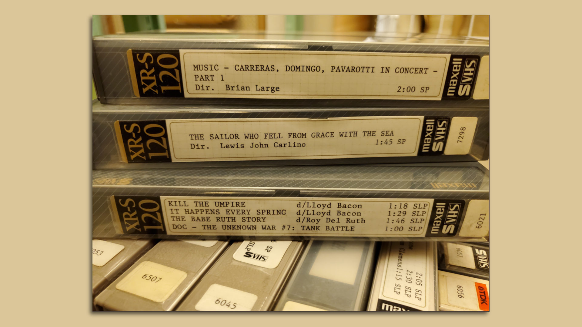 A stack of three VHS tapes, inside plastic covers, with words describing what's in each tape. The tapes are sitting on top several other tapes. 