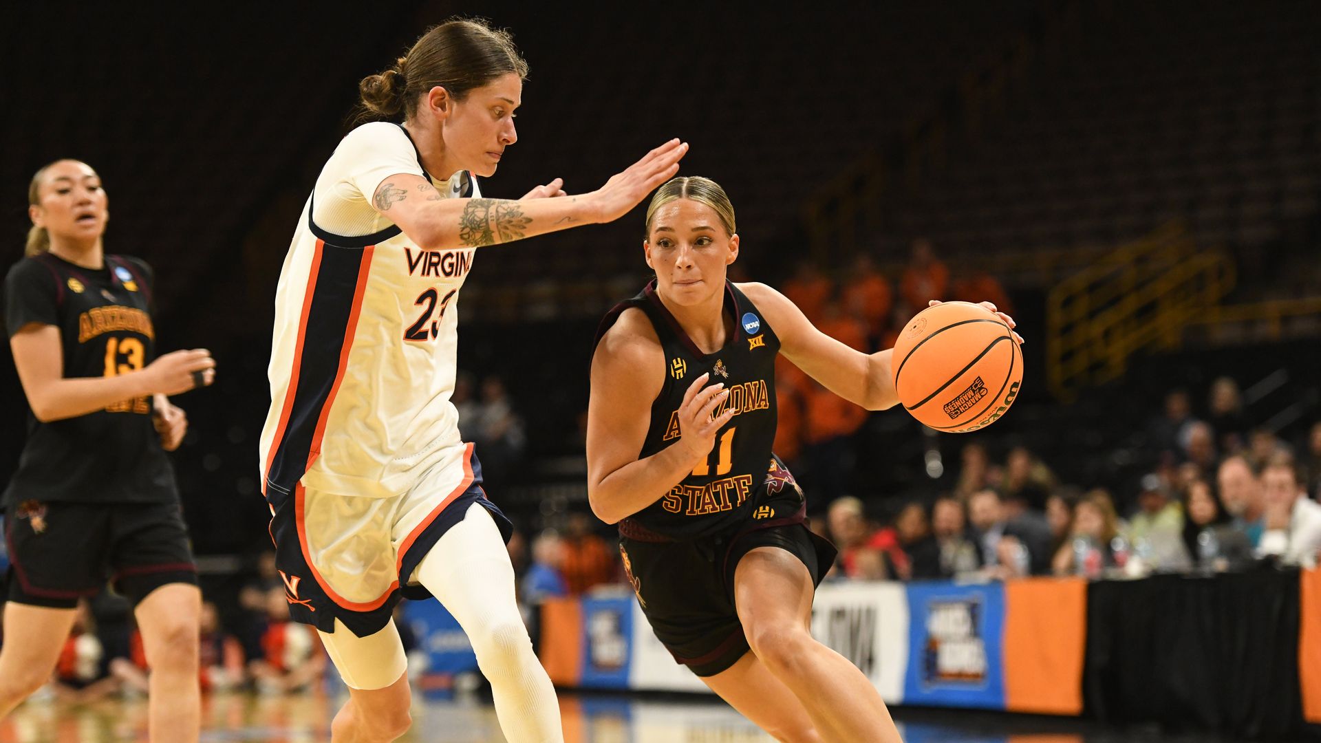 A woman in a black basketball uniform with the words "Arizona State" in crimson lettering a yellow "11" on the front drives with the ball in an arena against a defender in a white uniform with blue and orange stripes down the side. 