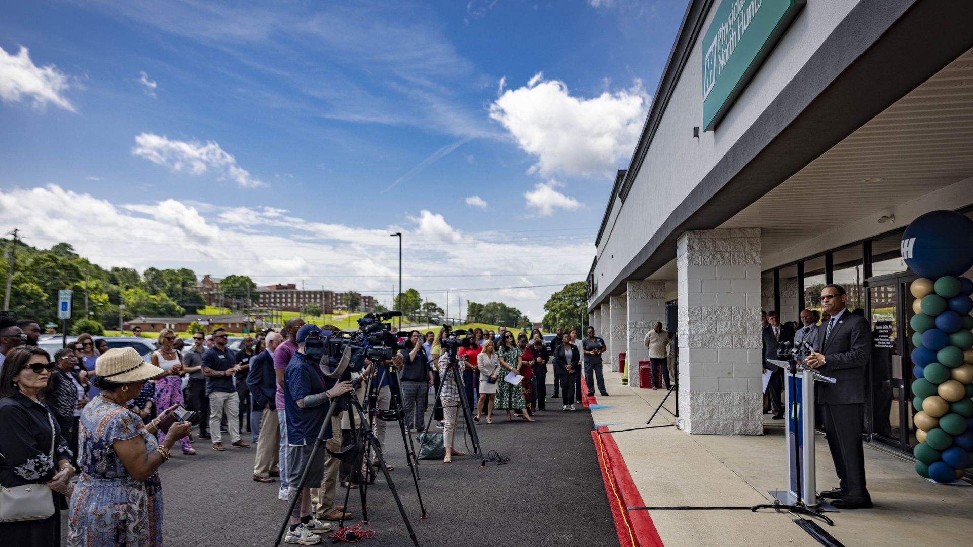 Outside a storefront, a crowd of journalists and attendees watches a speaker at a podium. Camera crews line the sidewalk; teal sign and balloon columns frame the entrance under a blue sky.