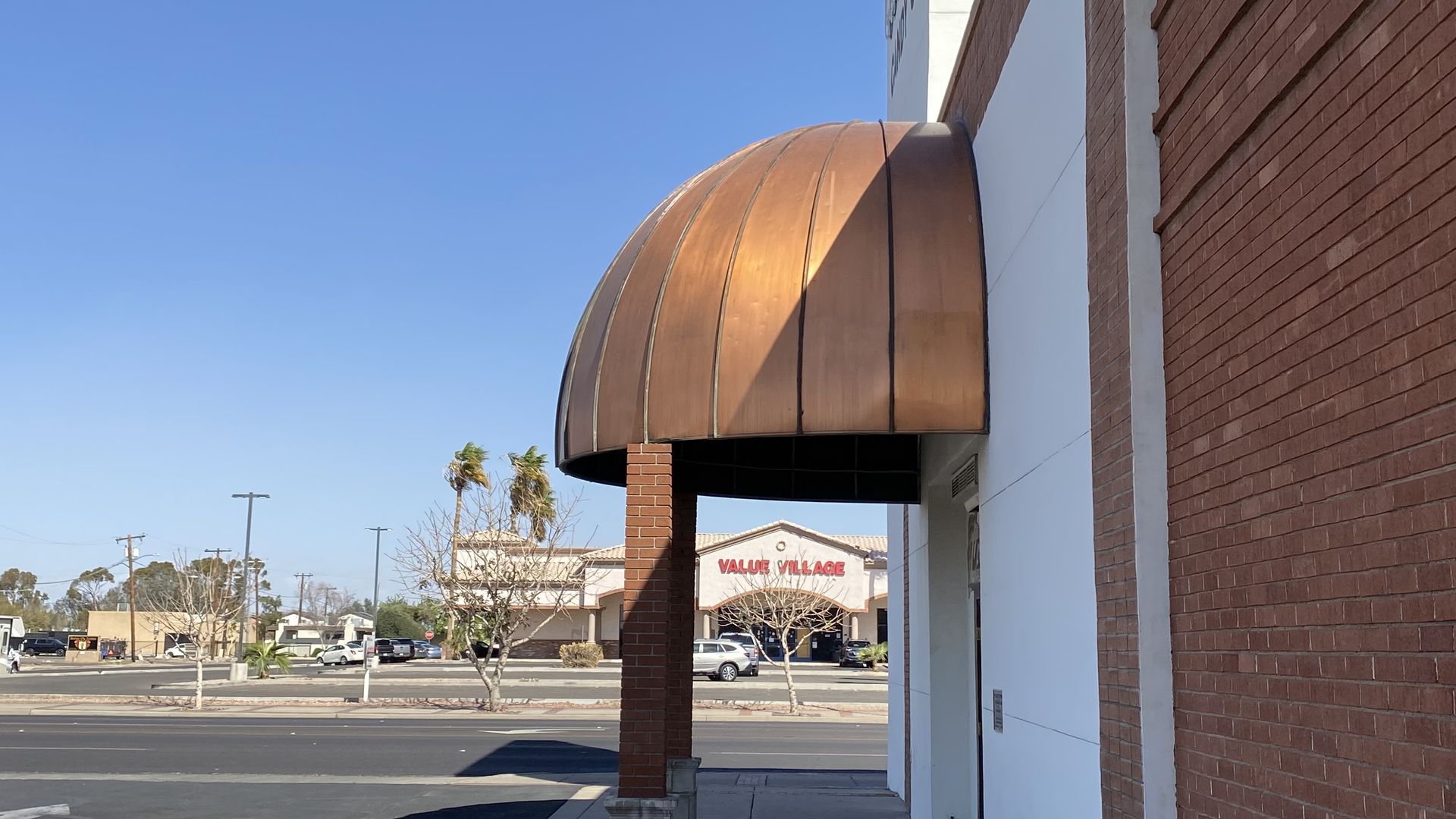 Street view showing a copper-domed awning attached to a red brick and white building, with a parking lot and store named Value Village in the background under a clear blue sky.