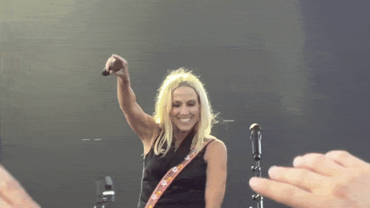 Blonde female guitarist on stage, wearing a black sleeveless top and a colorful guitar strap, smiles and raises her right arm while a crowd reaches up; a mic stand is nearby against a dark backdrop.