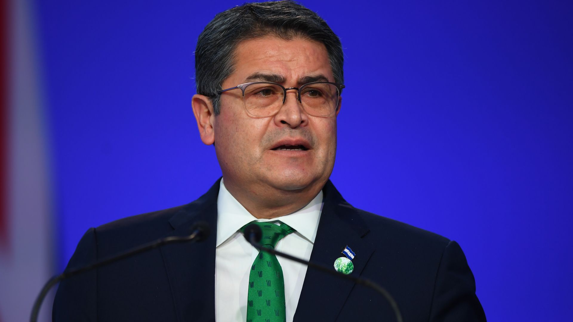 Ex-Honduran president Juan Orlando Hernández stands in front of a microphone while wearing a green tie and black suit. 