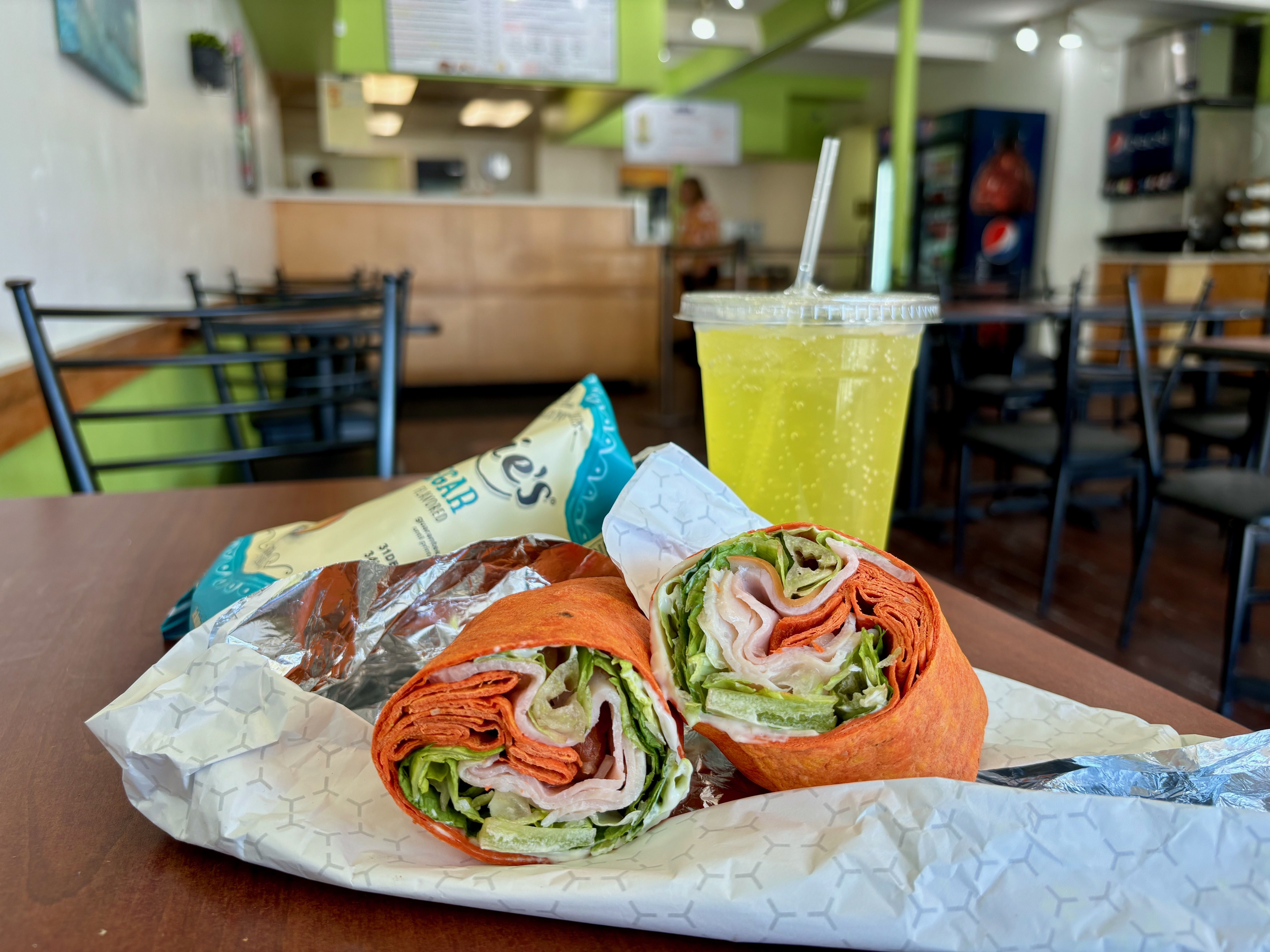A lunch of deli wrap, chips and drink on a table. 