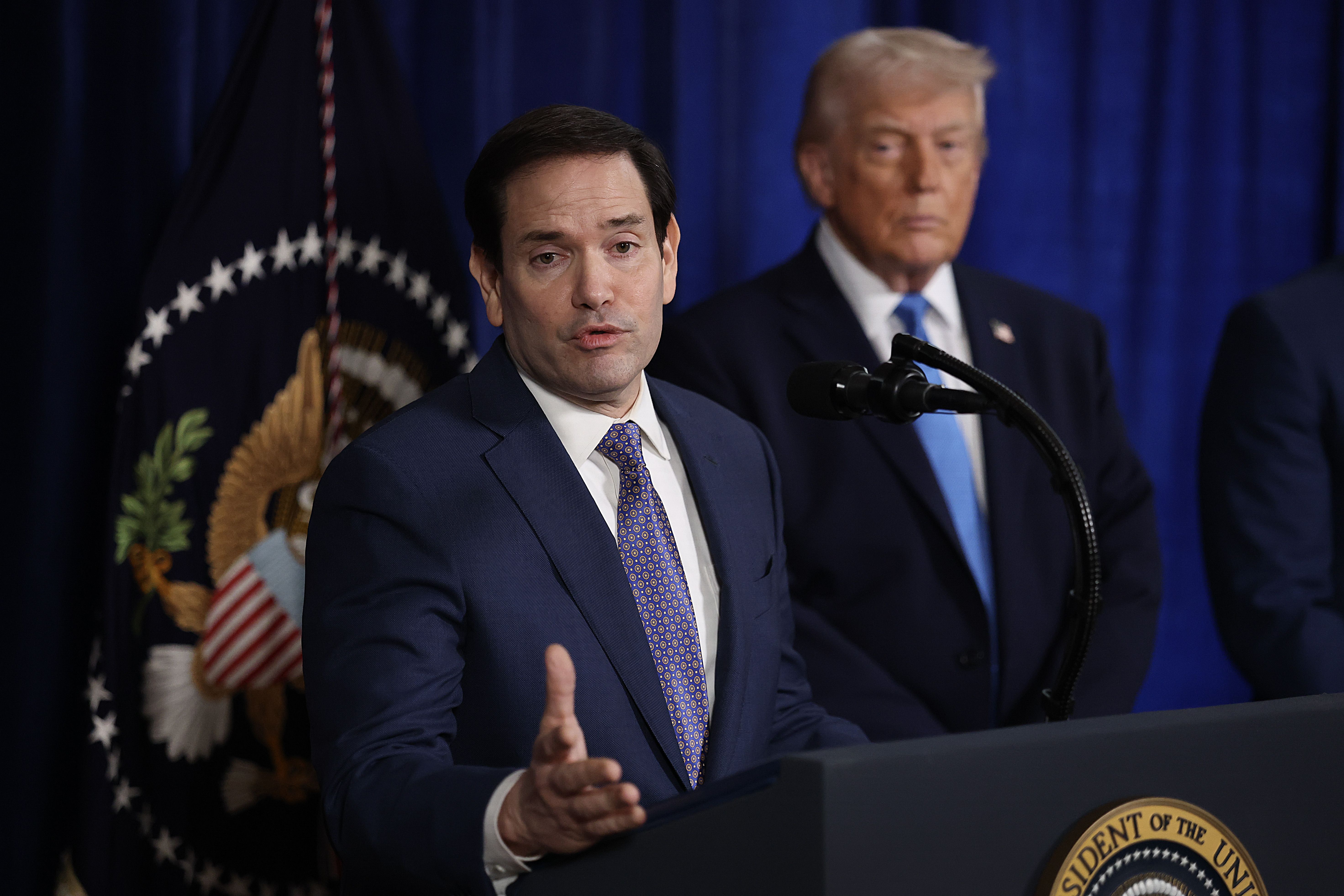 U.S. Secretary of State Marco Rubio at a podium with President Donald Trump listening beside him at a Mar-a-Lago press event.