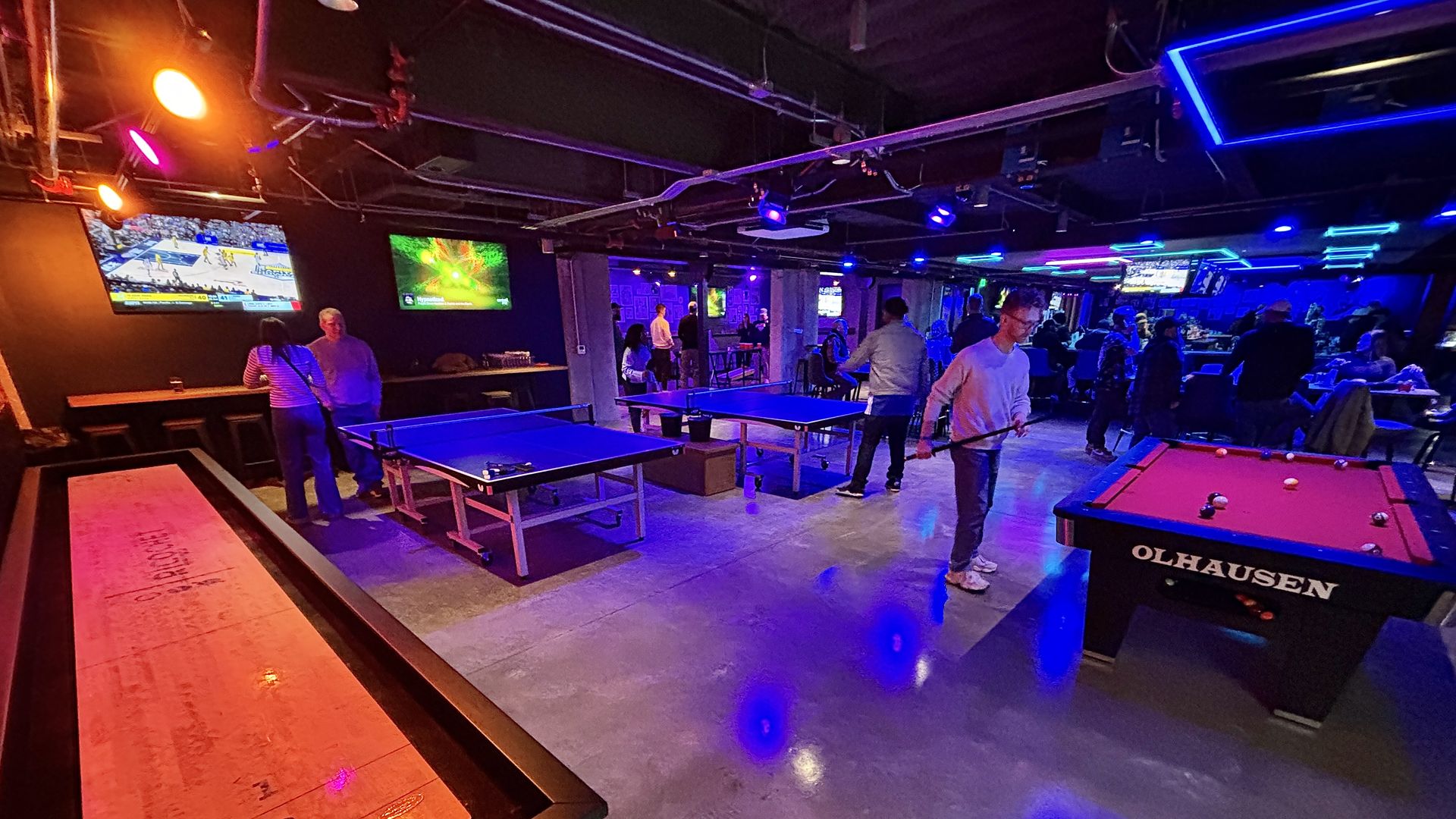 Indoor recreation area at Ricochet with blue-lit ping pong tables, a red pool table, people playing games and watching TV screens showing sports and entertainment.