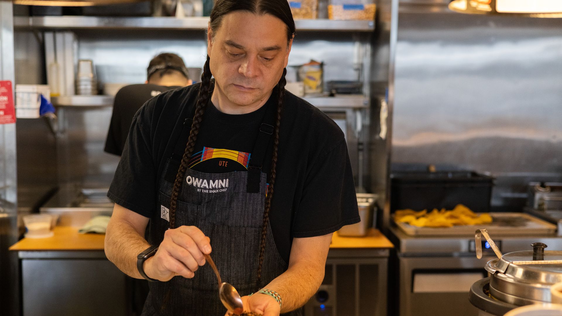 A man with olive skin and two long braids of dark hair holds a spoon in a kitchen as he cooks