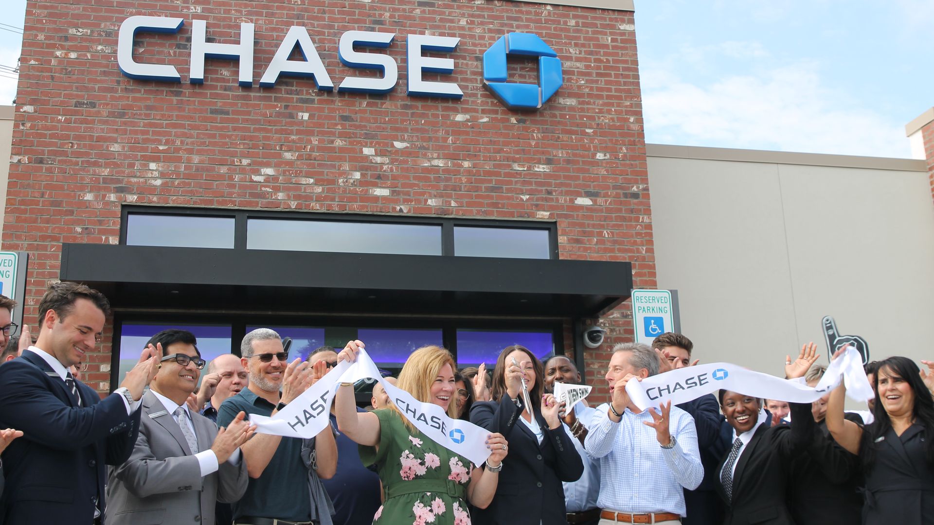 Group of people cheer and hold a white ribbon at a Chase bank branch ribbon-cutting event outside a brick building under a blue sky.