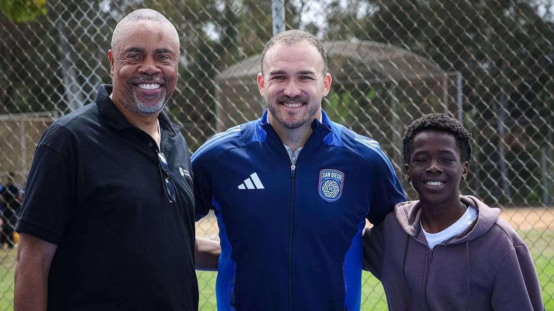 Three people smile for a photo on a sports field in front of a chain-link fence: an older man in a black polo, a man in a blue Adidas jacket with San Diego crest, and a boy in a purple hoodie.
