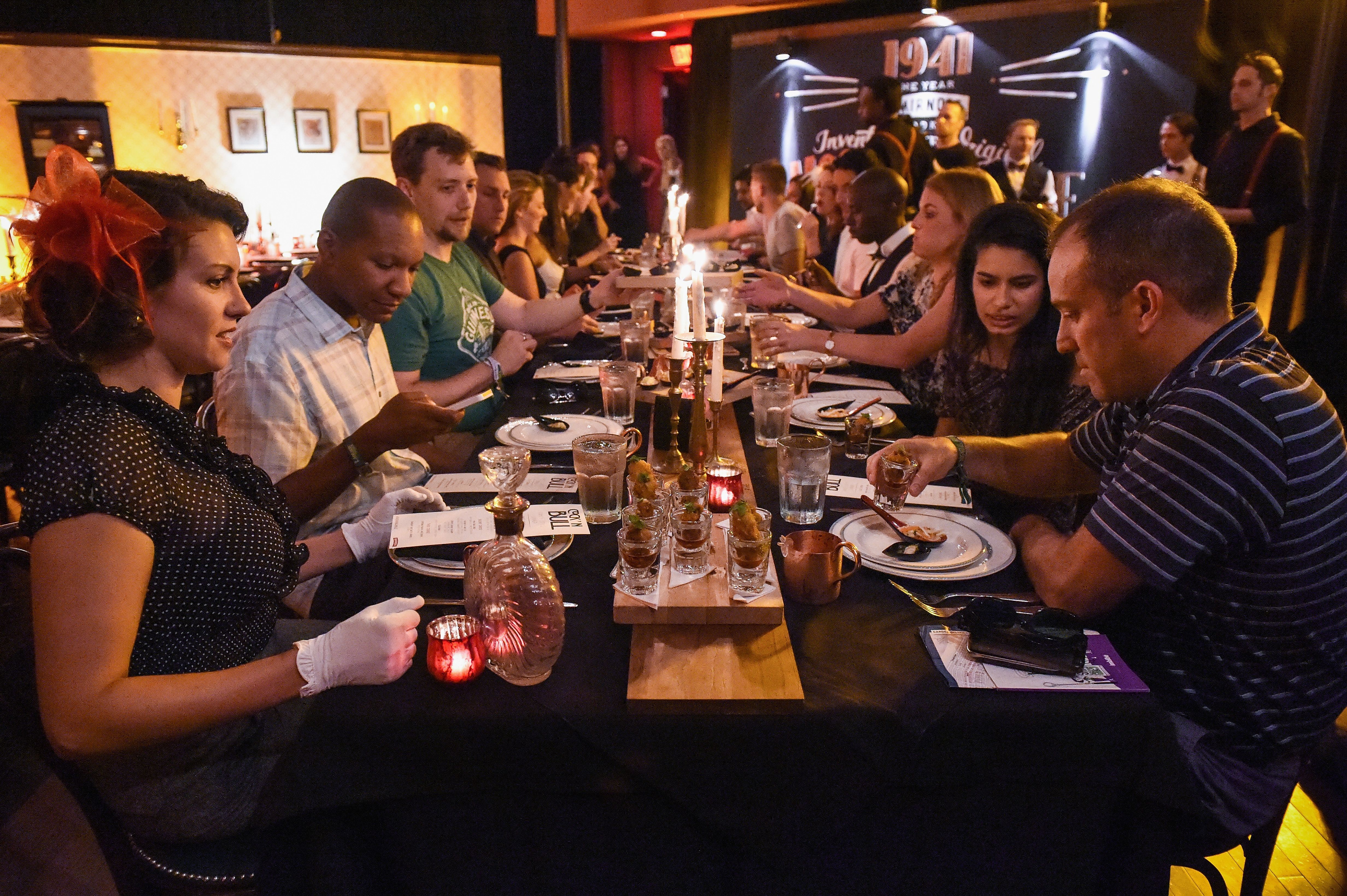 A view down a long dinner table with many guests seated at the table. They mingle and look at the menu cards placed on the plates in front of them.