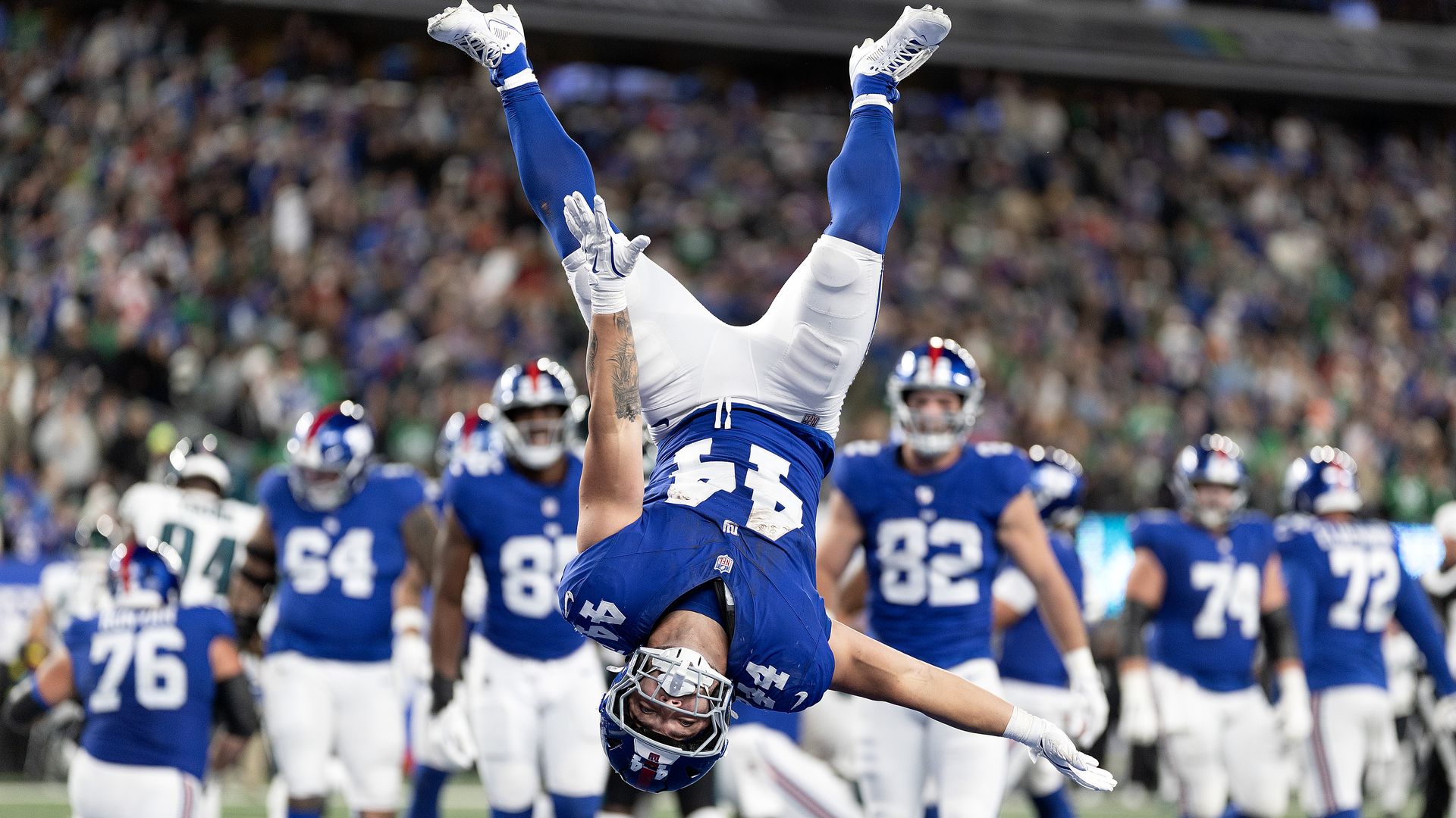 A football player in a blue jersey and white pants flips upside down in the air while celebrating in the end zone, while several teammates look on in the background. 