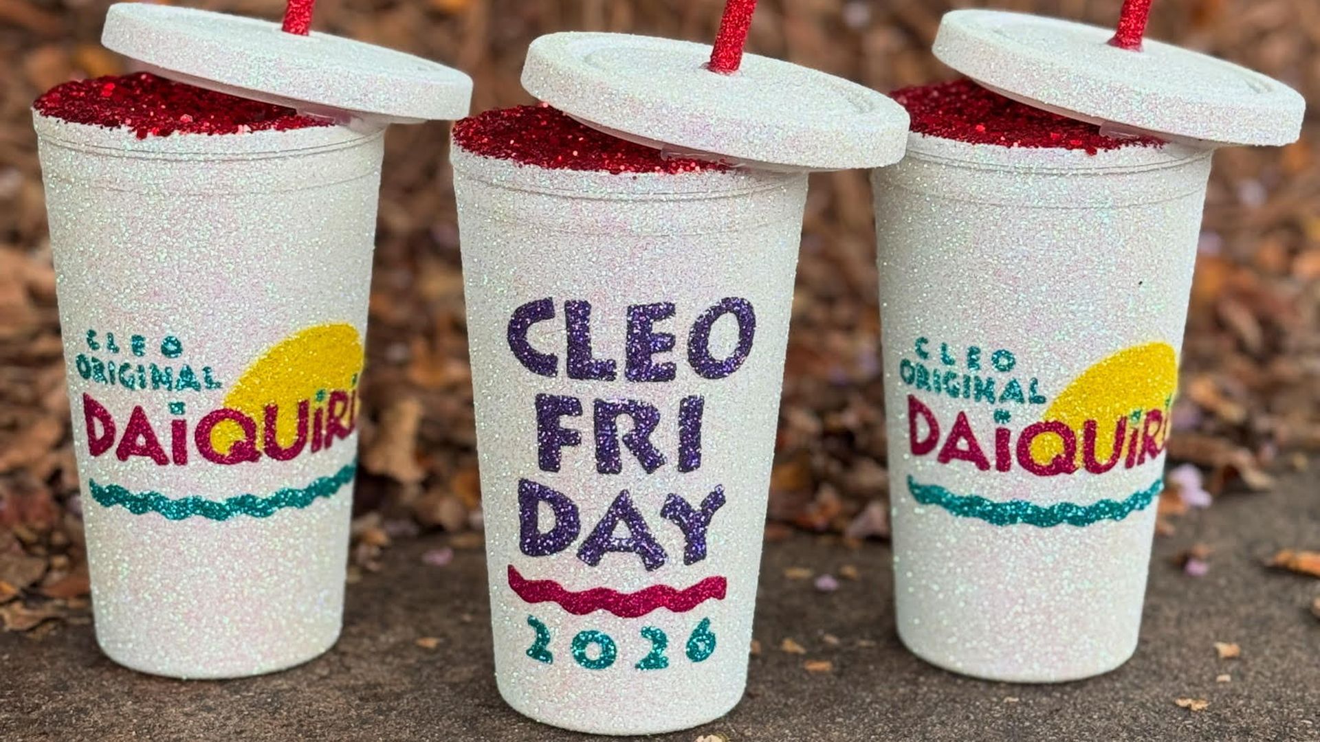 Three glittery cups with red straws and lids, two labeled "CLEO ORIGINAL DAIQUIRI" with a yellow and red design, and one labeled "CLEO FRIDAY 2026" in purple and teal text.