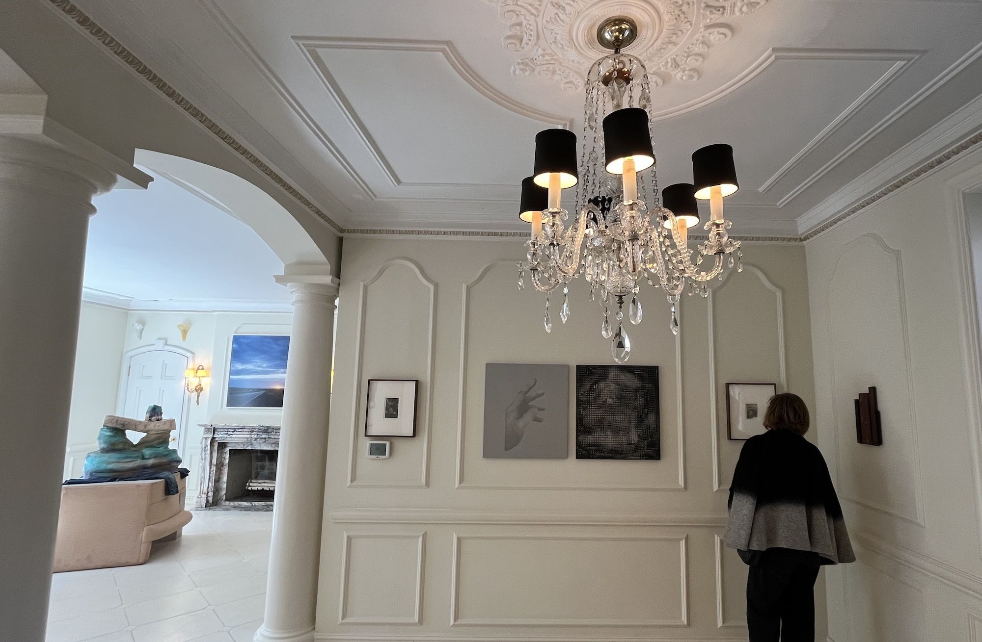 Elegant cream room with ornate plaster ceiling, crystal chandelier, and paneled walls. A person in a gray cape stands by framed art; arched doorway and marble fireplace visible in the background.