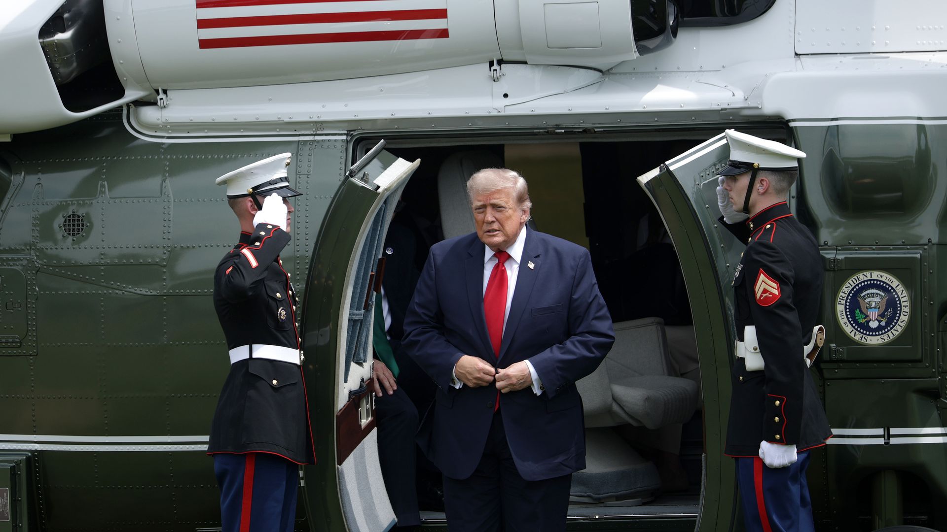 President Trump  in a dark suit and red tie steps from a white and green helicopter, flanked by two Marines saluting in dress uniform, by the open door on a grassy field; a flag decal is on the fuselage.