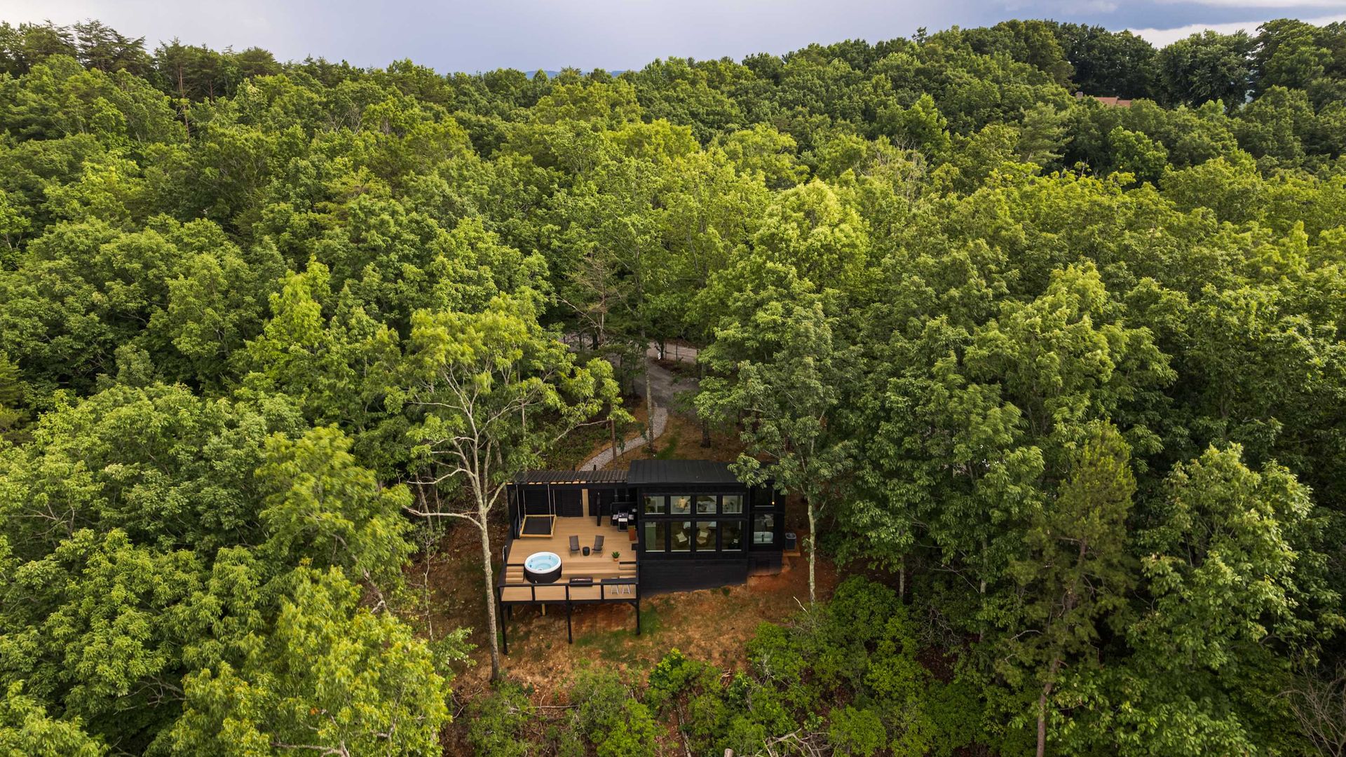 A black modern mountain cabin studio with a hot tub deck sits in a dense forest 