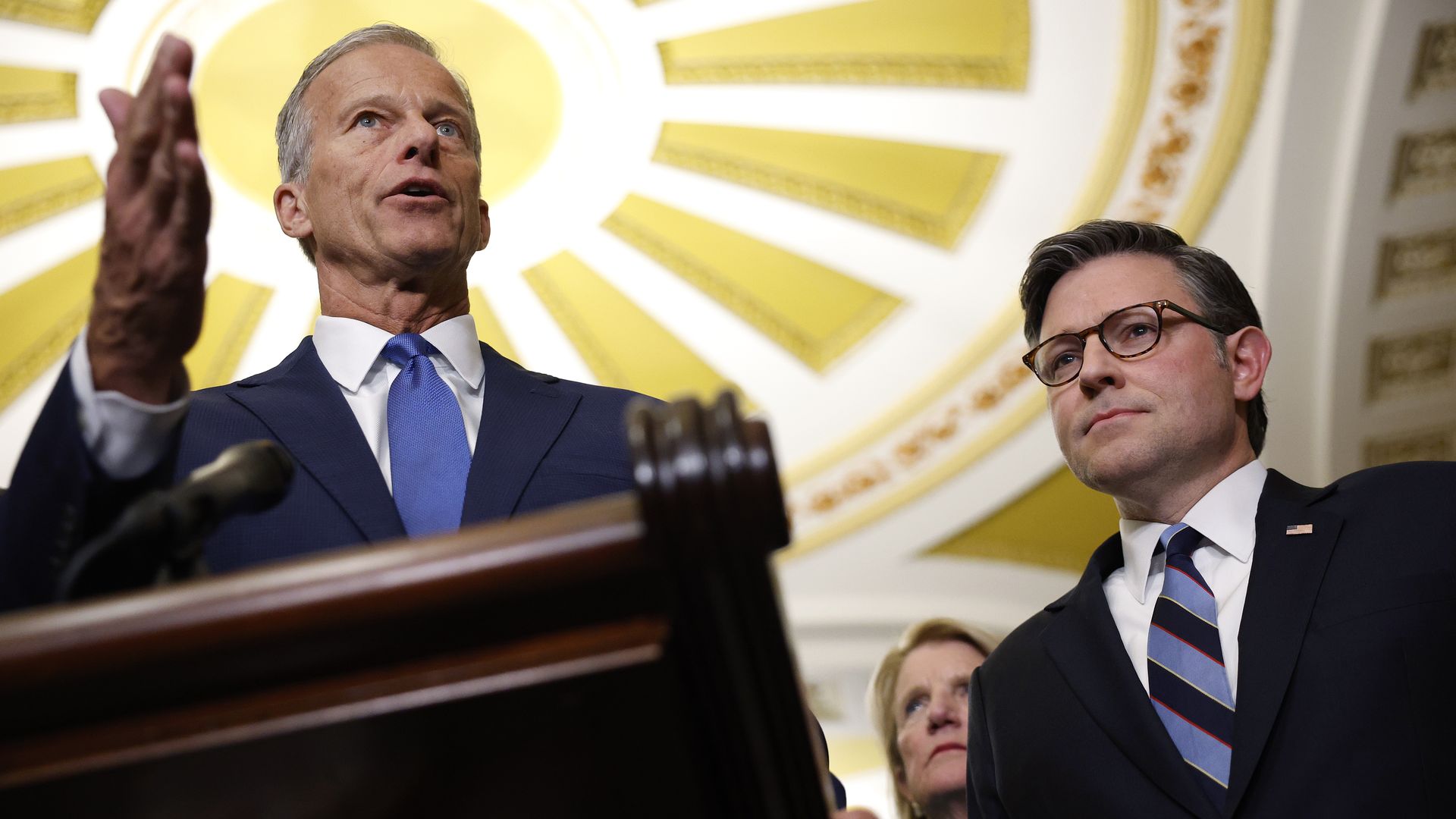 Two men in suits speak at a podium under a large yellow and white decorative ceiling, with a woman standing behind them listening attentively.