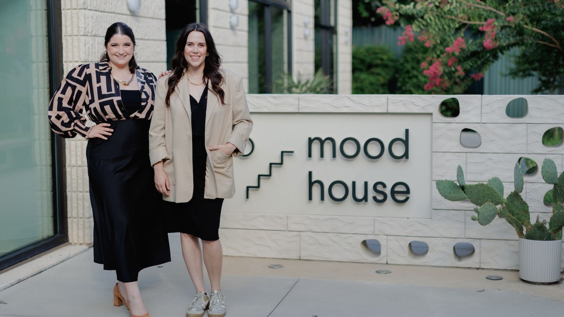 Two women stand outside a modern building beside a sign reading "mood house." Left wears a black dress with a patterned beige top; right wears a black dress with a beige blazer. A cactus sits to the right.