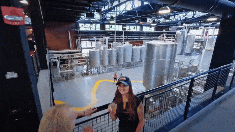 Interior of a brewery with large silver fermentation tanks and pipelines. Two women in the foreground are tasting beer; one wears a black cap and shirt, the other reaches out holding a glass.
