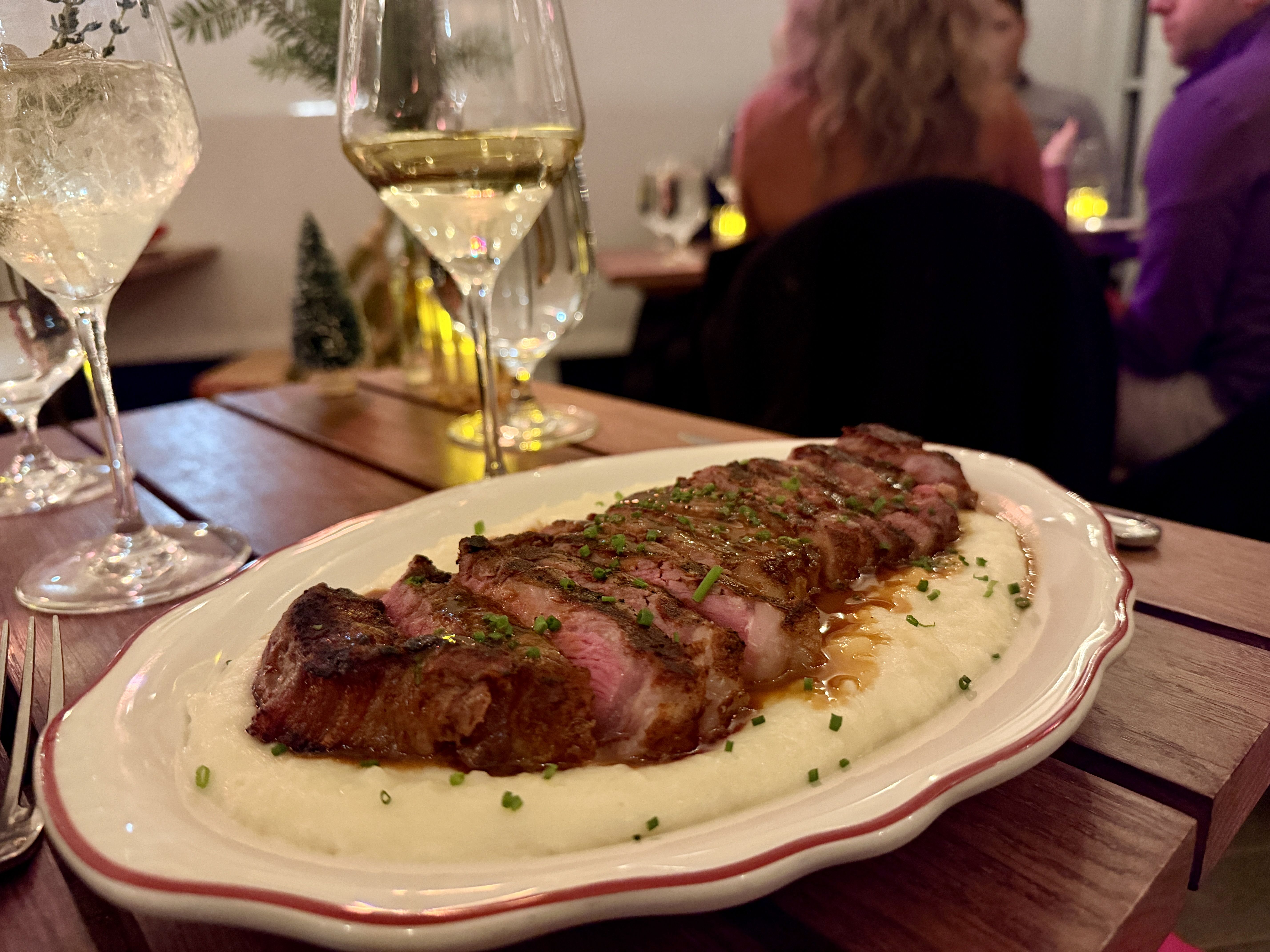 Sliced medium-rare steak garnished with chopped chives, served on creamy white mashed potatoes on a white plate with a red rim, in a cozy restaurant setting with wine glasses nearby.