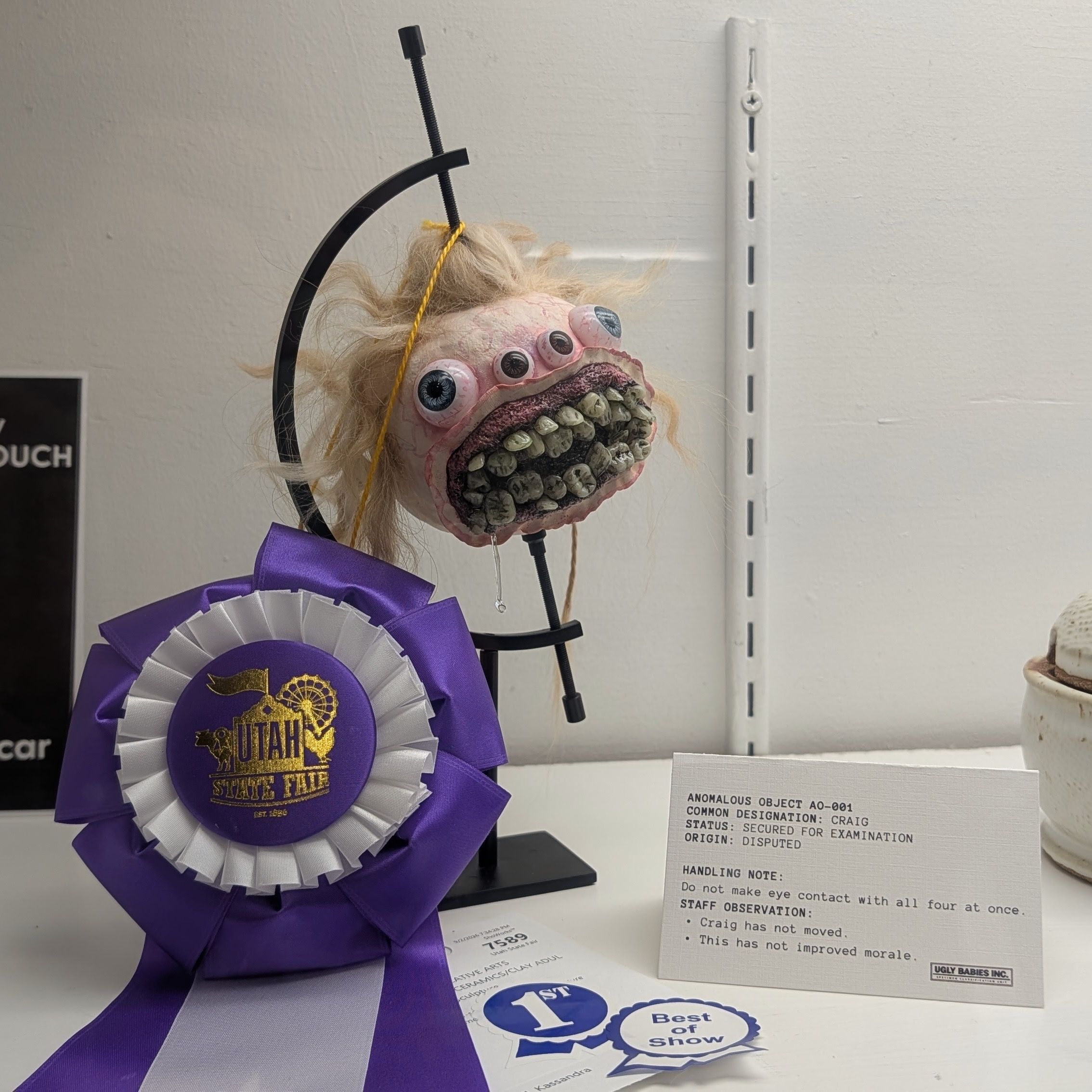Grotesque puppet head with many eyes and a toothy mouth, suspended by a black arc frame; a purple Utah State Fair ribbon rests in front against a light wall.
