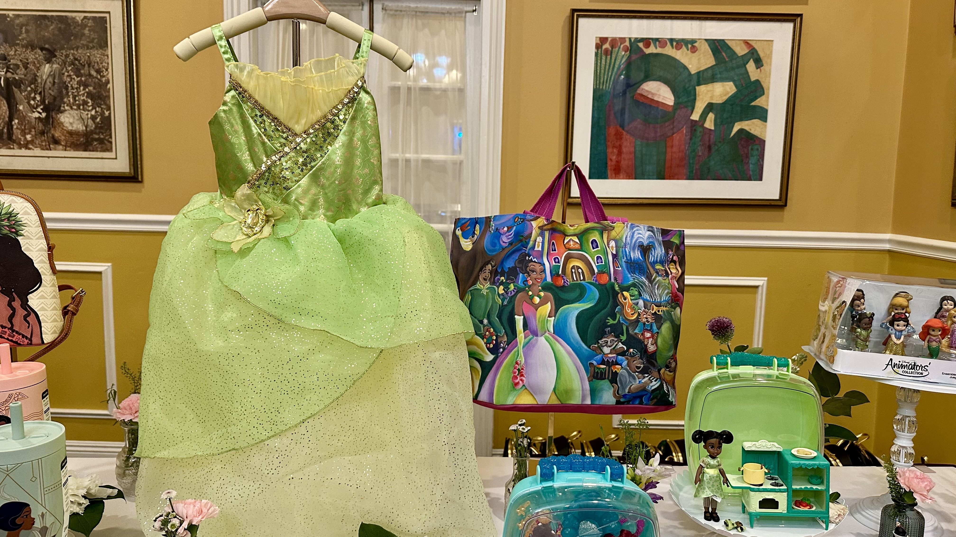 Green and yellow sparkly dress on a hanger, colorful tote bag with cartoon characters, small doll with green outfit and kitchen playset, figurines set on a yellow-walled room table.