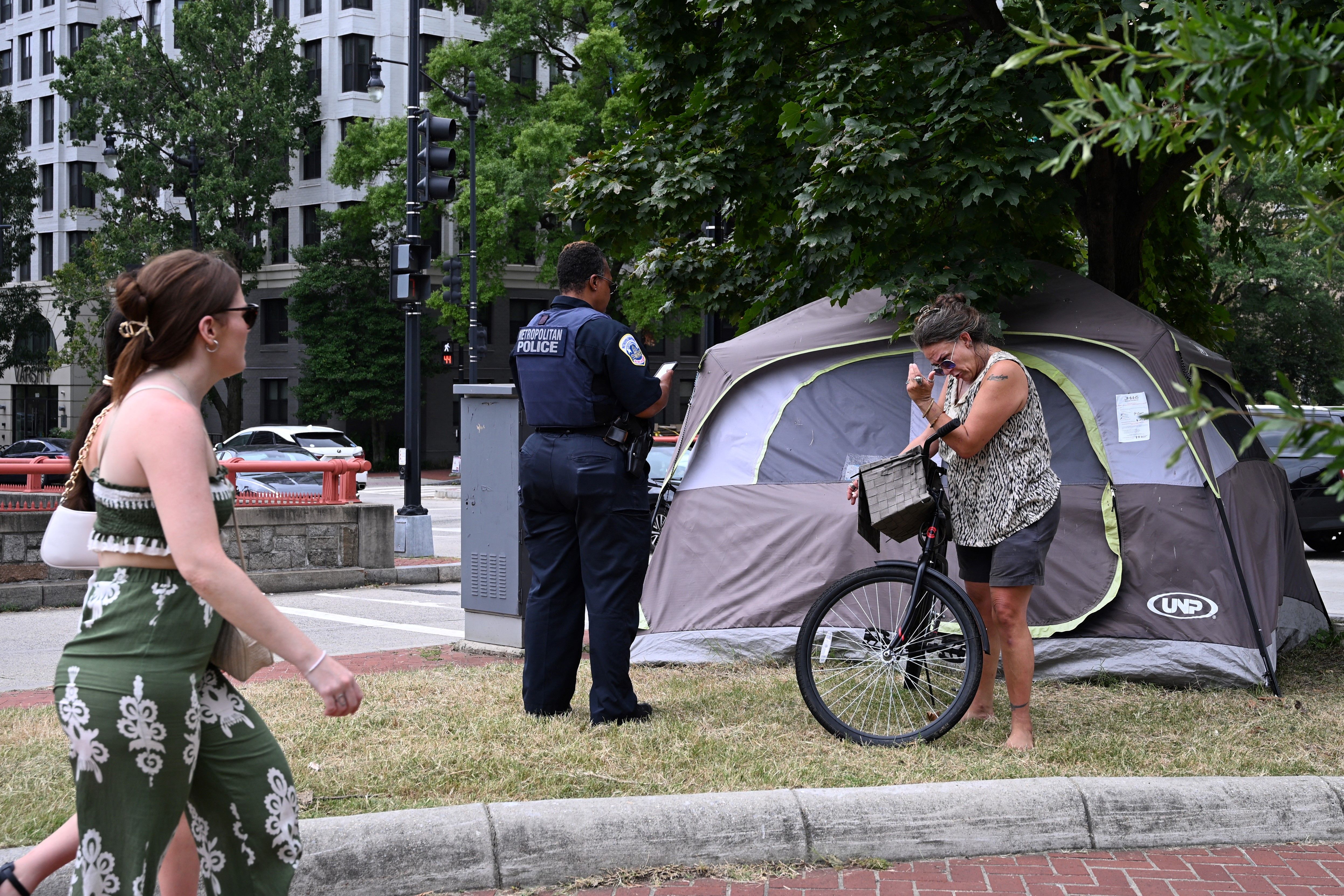 A Metropolitan Police officer writes notes near a gray and green tent while a barefoot woman with a bicycle looks down, as a woman in green patterned pants walks by on a city sidewalk.