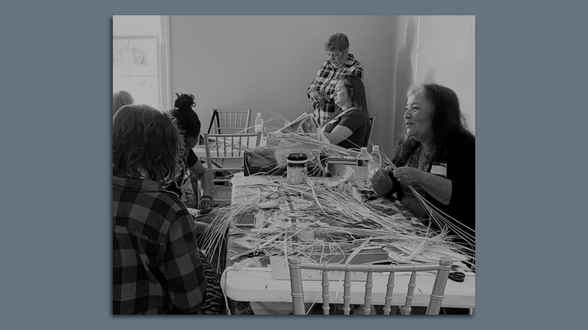 Several people gather around a large white table to weave baskets together. Janie Luster is seen at right.