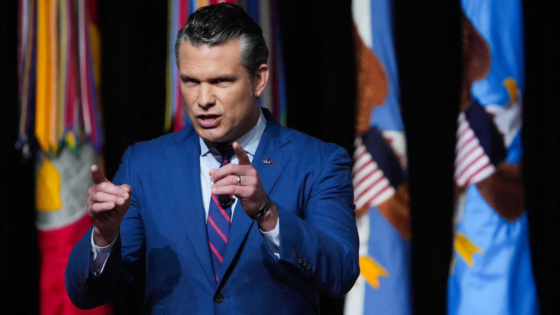 Pete Hegseth points with both hands as he speaks on a stage in front of several flags wearing a dark blue suit. 