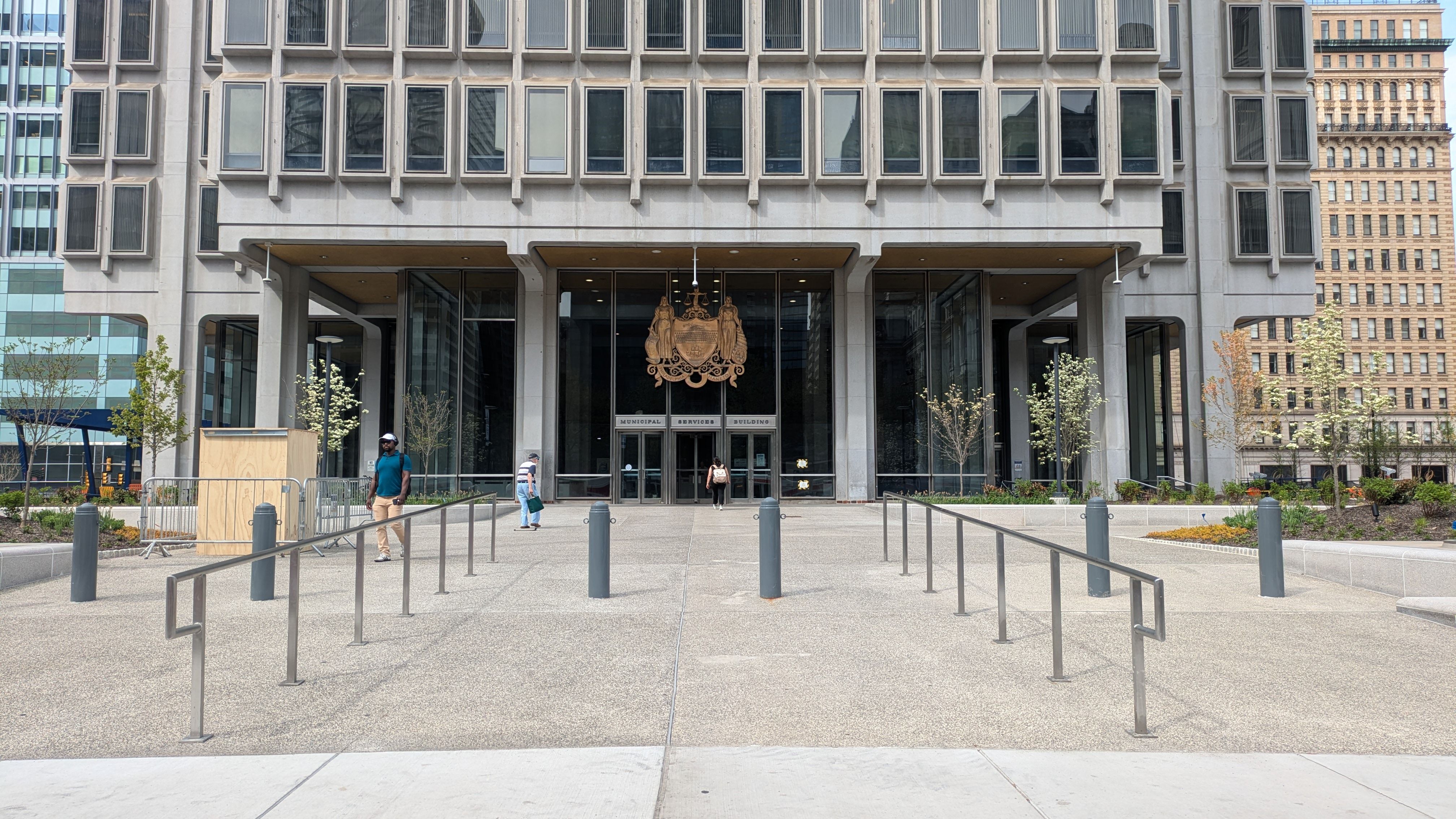 Front of a large concrete municipal building with a glass entry and a decorative crest above the doors. A plaza with rails leads to the entrance as a few people walk toward it, with trees around.