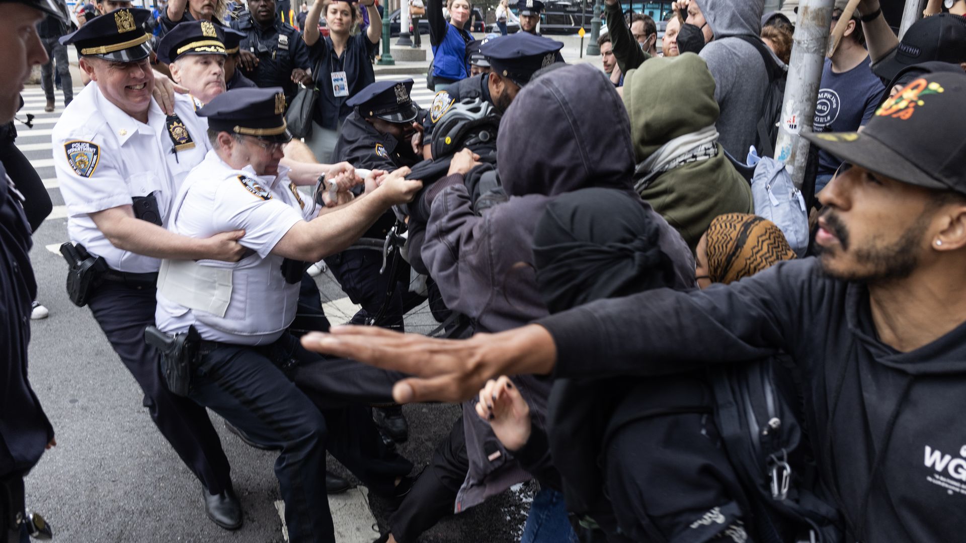 A street protest scene showing a tense confrontation between police officers and protesters. Several police officers, dressed in uniform, are attempting to control or detain individuals, while the protesters appear to be pushing back. The crowd is densely packed, with some people holding up cameras 