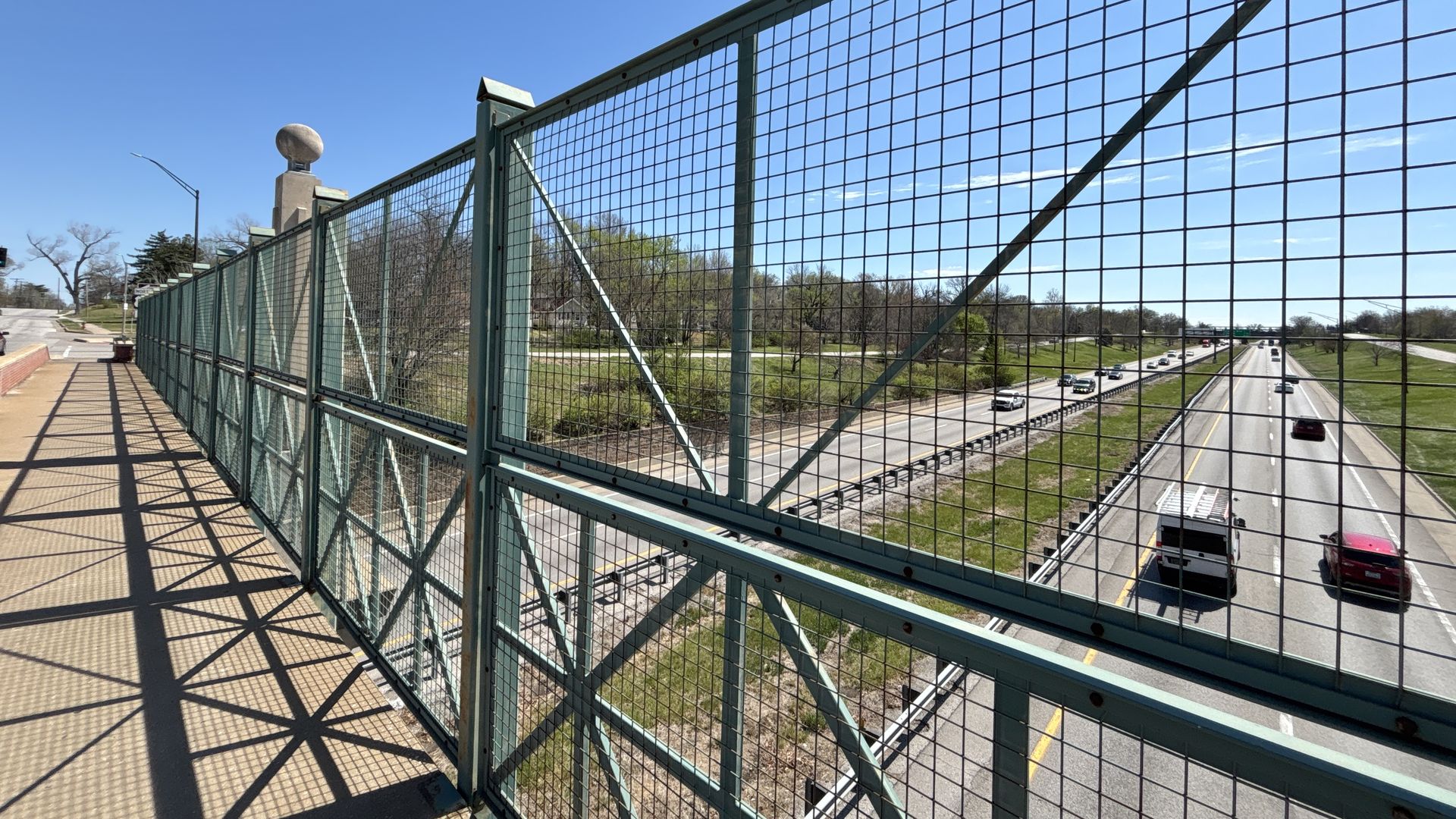 Pedestrian bridge with a tall green metal grid fence; walkway casts lattice shadows. Below is a freeway with cars, grassy medians, and trees under a clear blue sky.