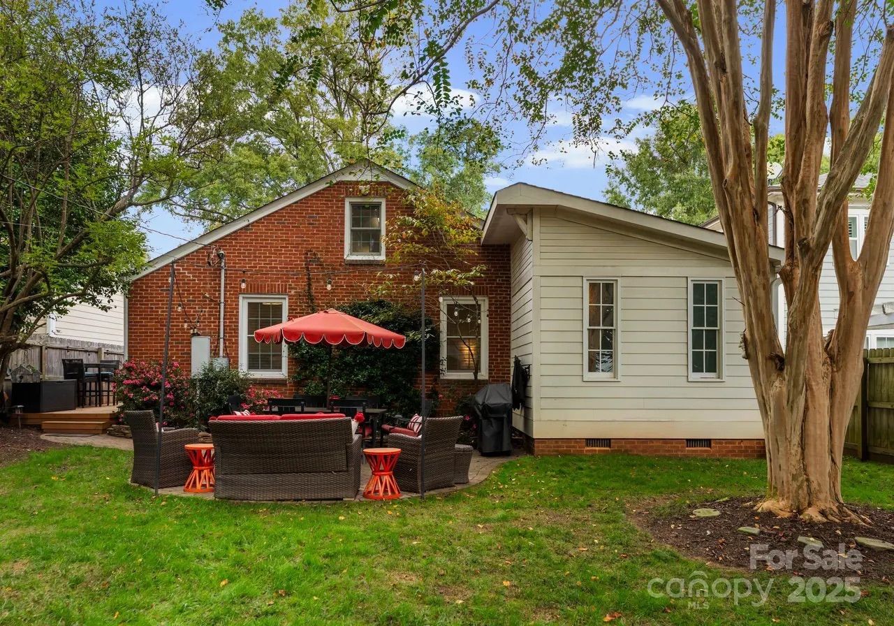 Backyard with green grass, patio furniture with red cushions and umbrella, brick and white siding house walls, trees, and a barbecue grill. "For Sale canopy 2025" watermark.