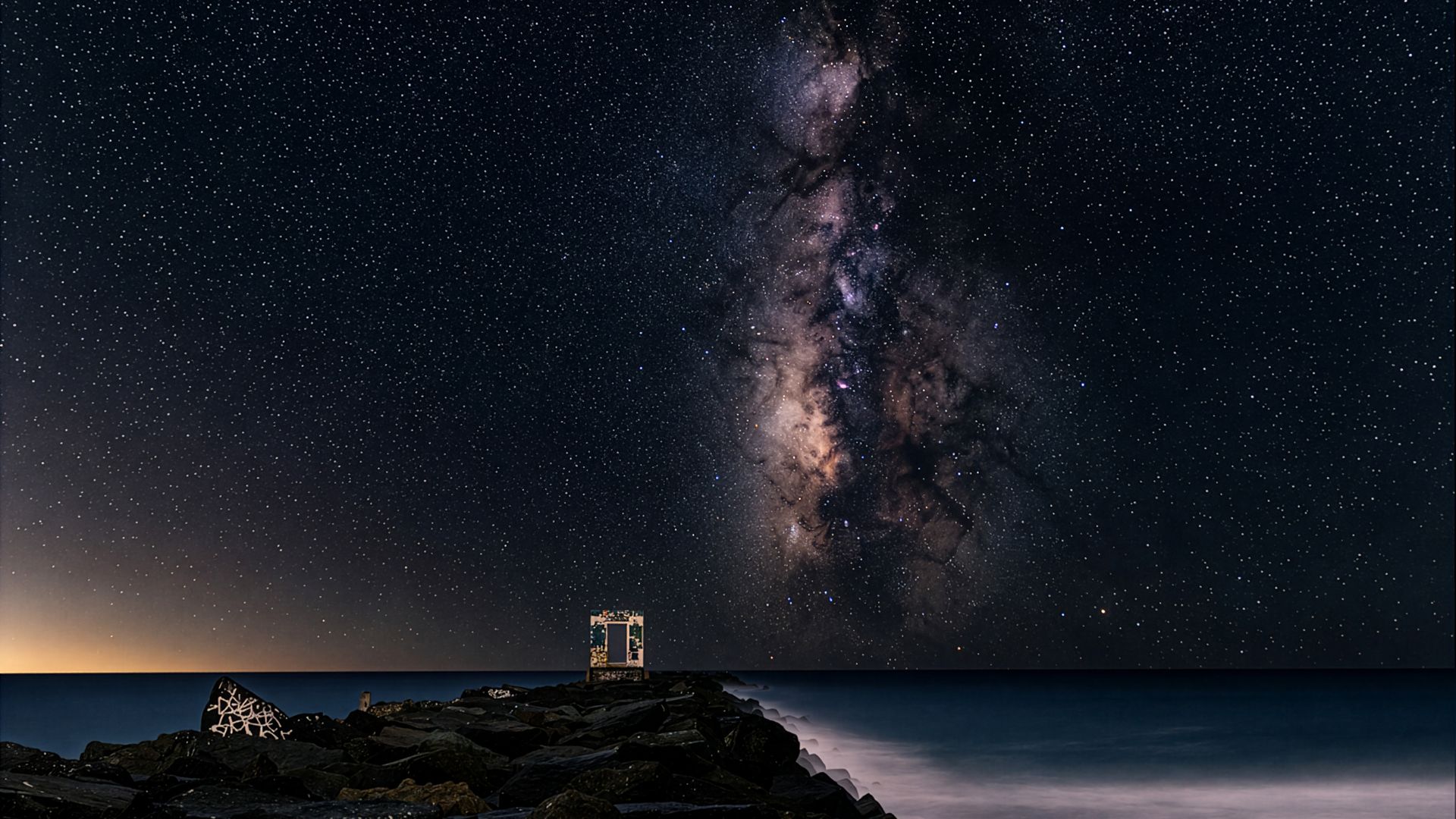 Night sky filled with stars and the Milky Way over a dark rocky shoreline; a small rectangular frame sits on the rocks by the water, with a faint orange glow on the horizon.