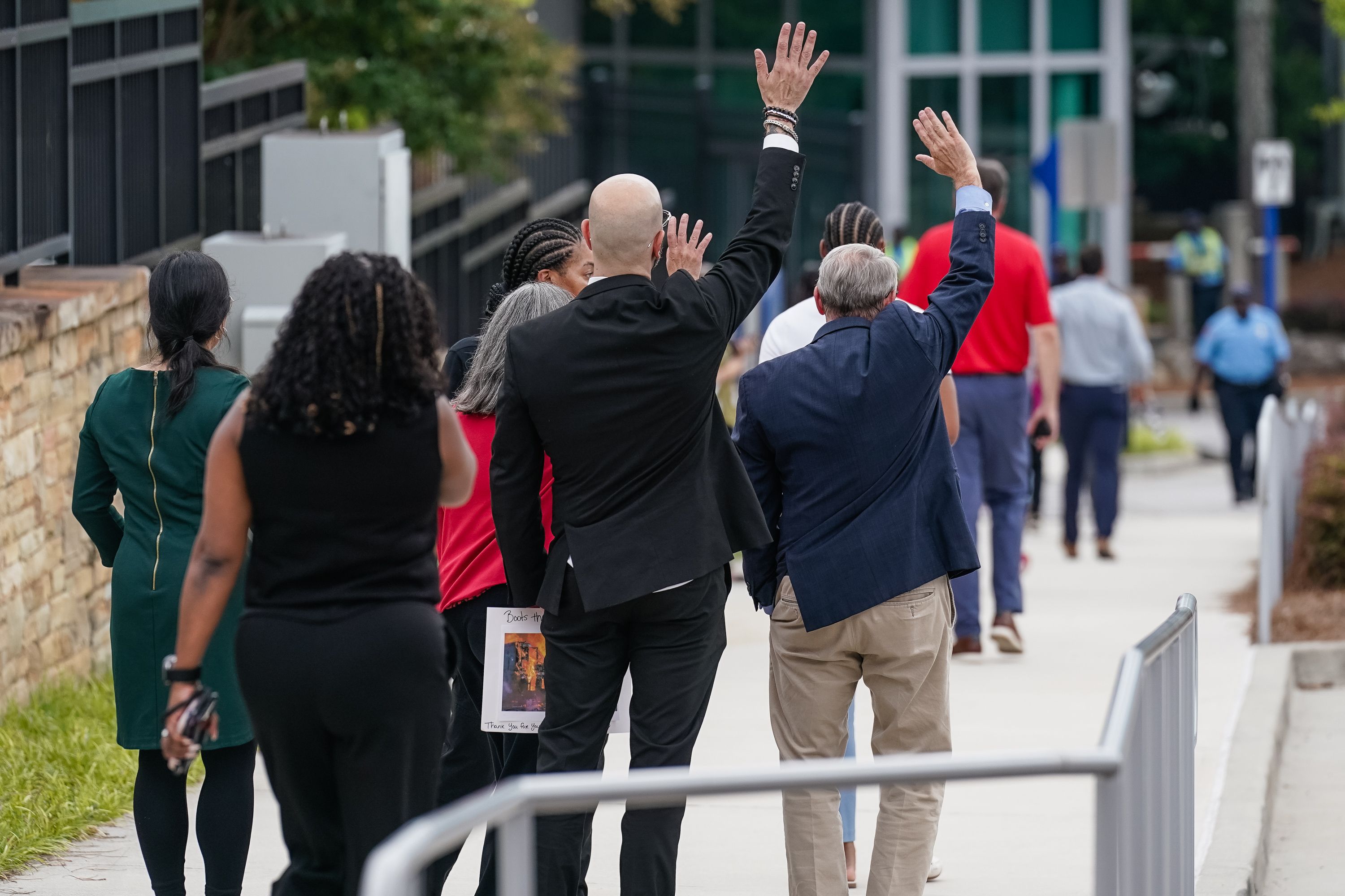 Group of diverse people, some in business attire, walking away on a sidewalk; two men in front wave their hands, one holding a paper; urban background with greenery and buildings.