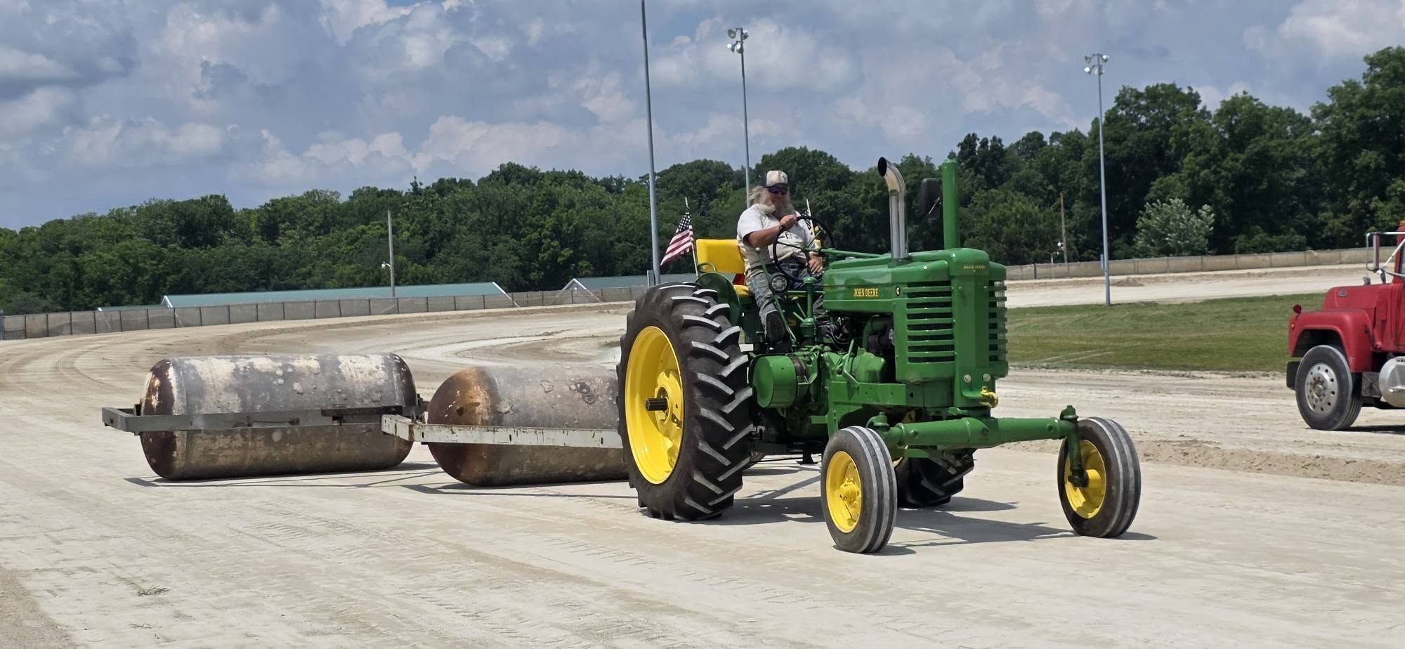 Green John Deere tractor with yellow wheels pulls two large road rollers along a dirt track; a man in a cap drives, an American flag waves, with trees and a red truck in the background.