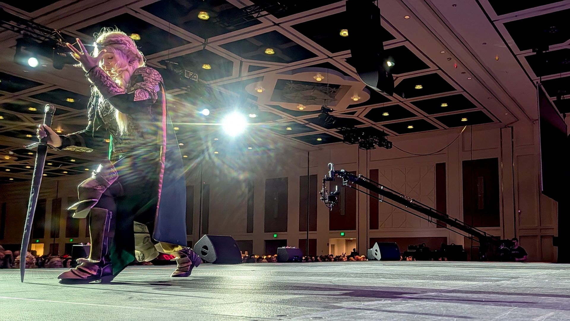 On stage, a performer with long blond hair in a purple-black costume grips a tall sword and microphone, under bright lights in a large venue with an audience watching.