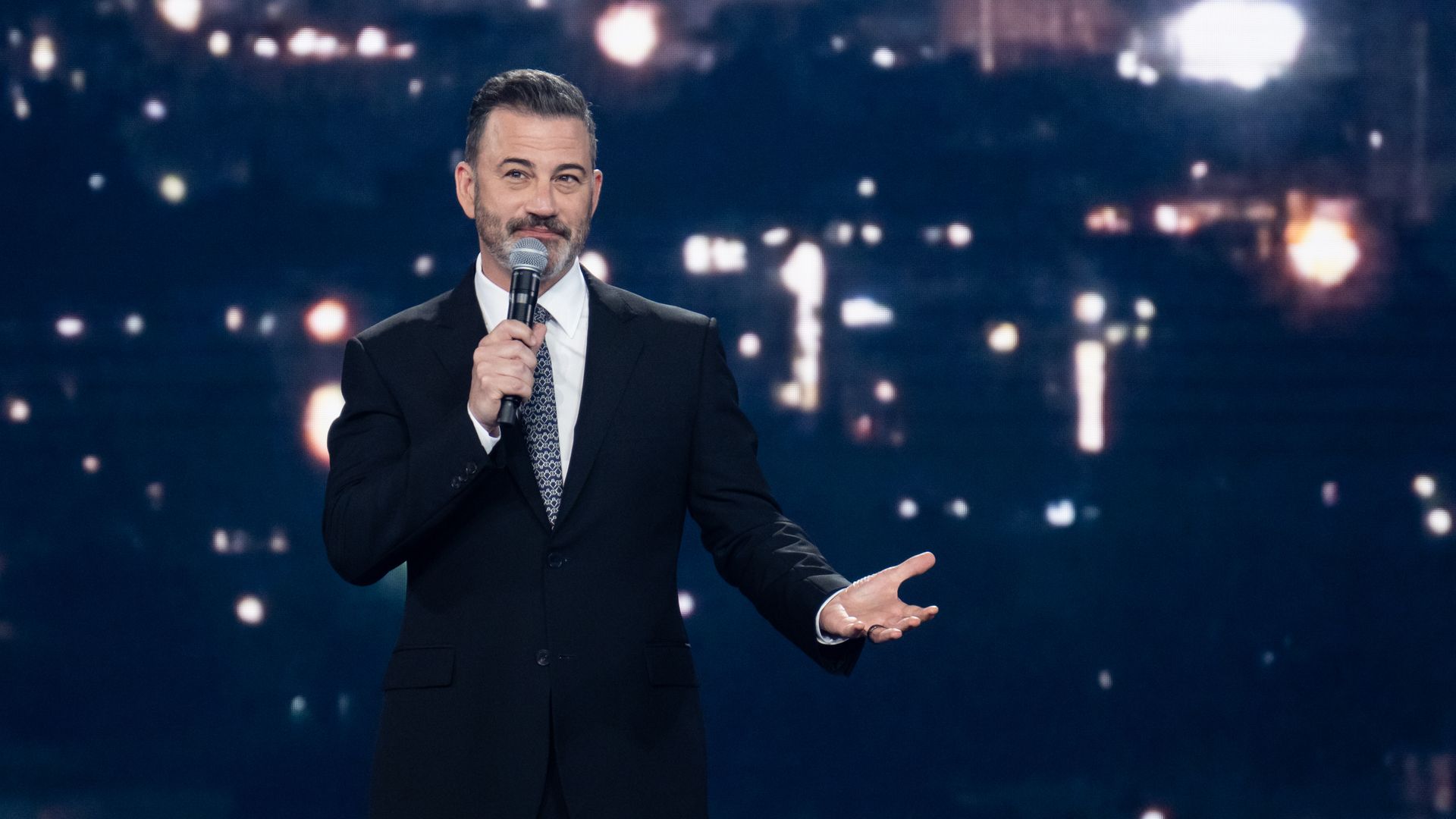Talk show host Jimmy Kimmel, wearing a black suit, white shirt and black and white tie, gestures in front of a city-scape screen.