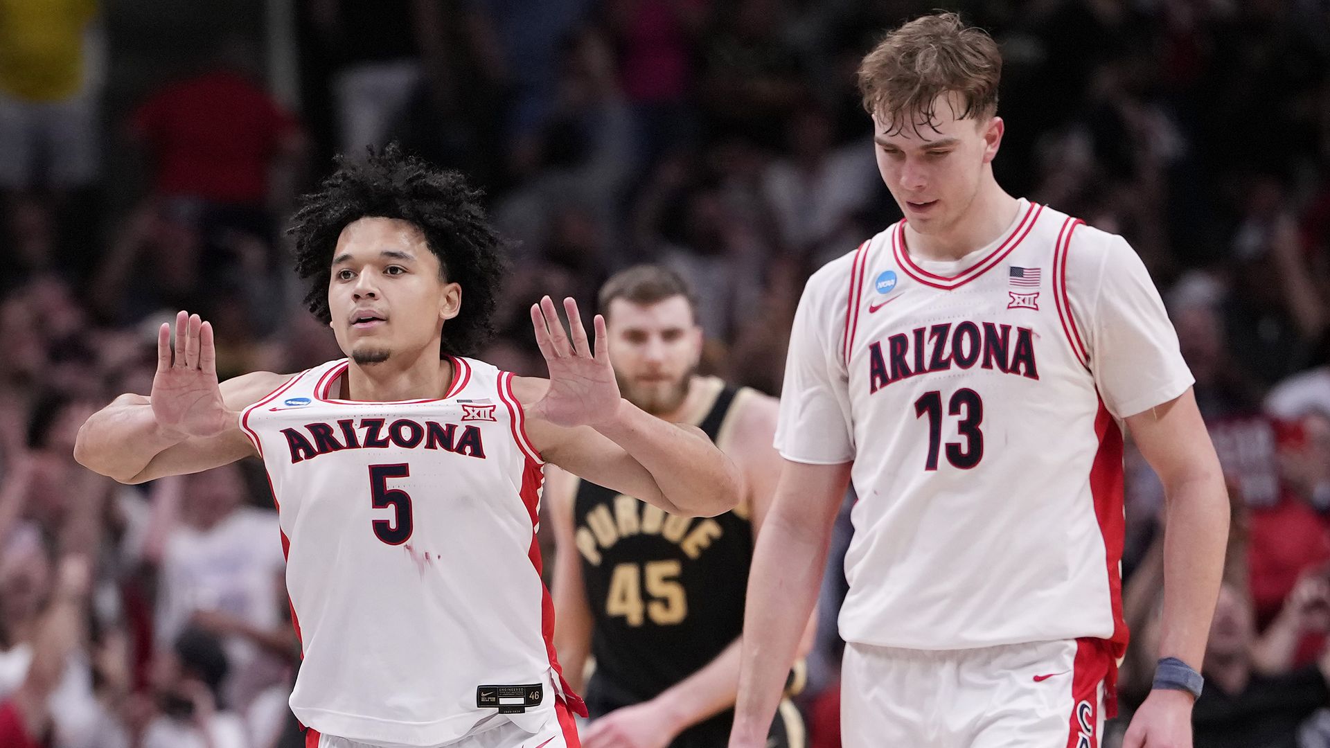 Two basketball players in white jerseys with "Arizona" on the front in dark blue lettering walk on the court; one of them is using his thumbs to pull the word "Arizona' forward. An opponent in a black uniform that reads "Purdue" walks in the background.