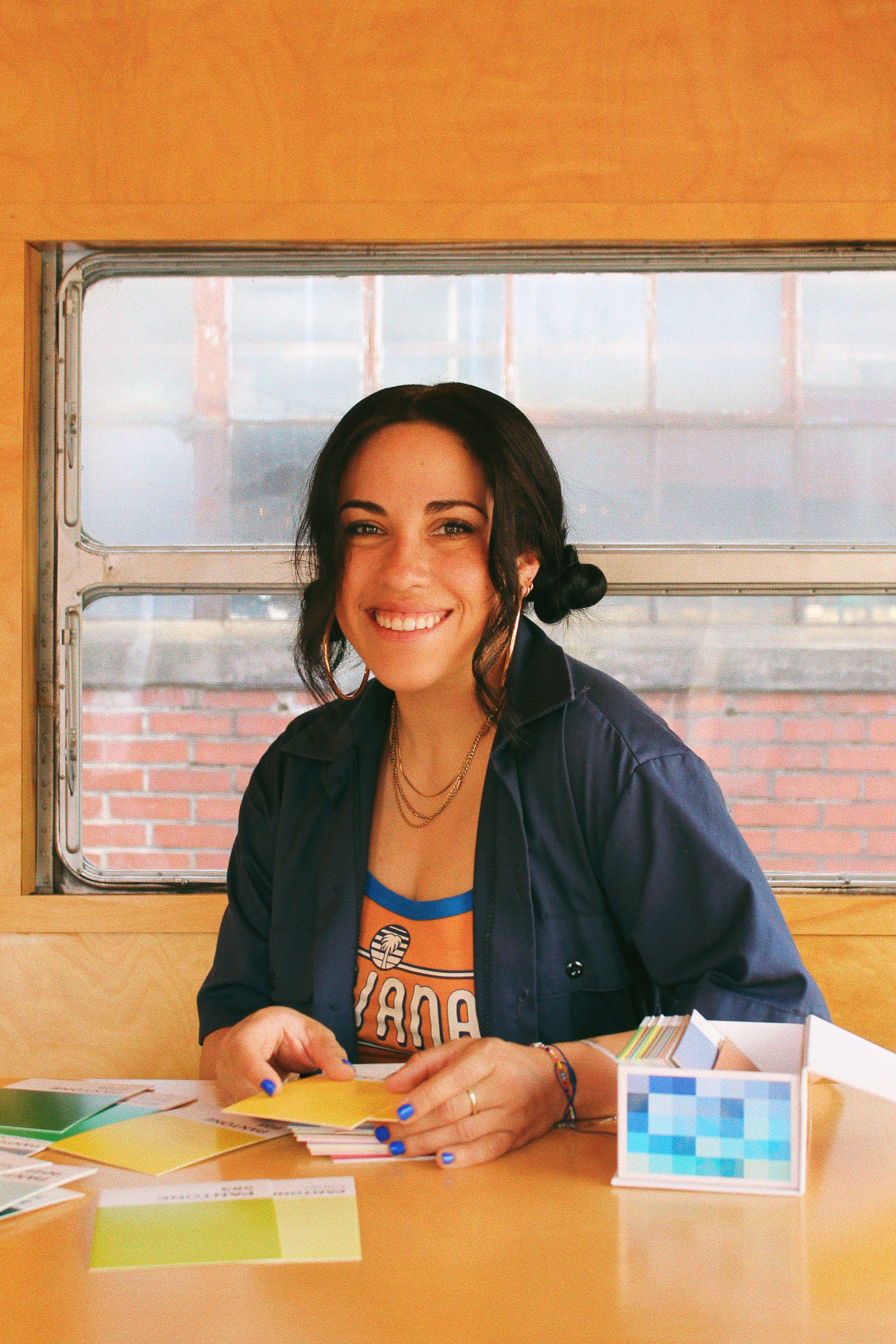 Smiling woman with dark hair and blue nail polish sits at a wooden table inside a yellow room near a window, sorting through colorful paint swatches and cards.