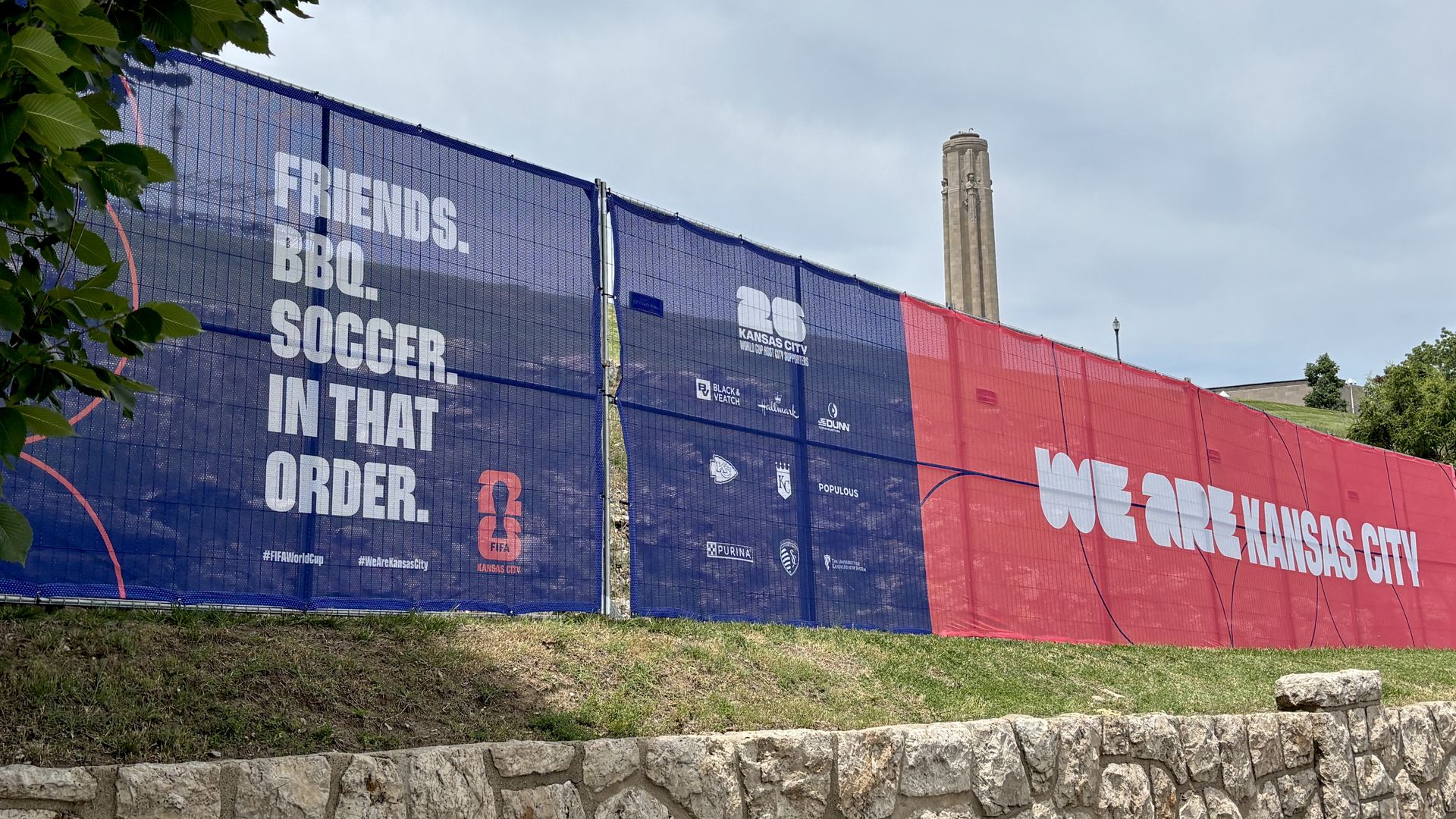 Blue and red construction fence banners display "FRIENDS. BBQ. SOCCER. IN THAT ORDER." with KC logos, a grassy slope, a stone wall, and a tall memorial tower in the distance.