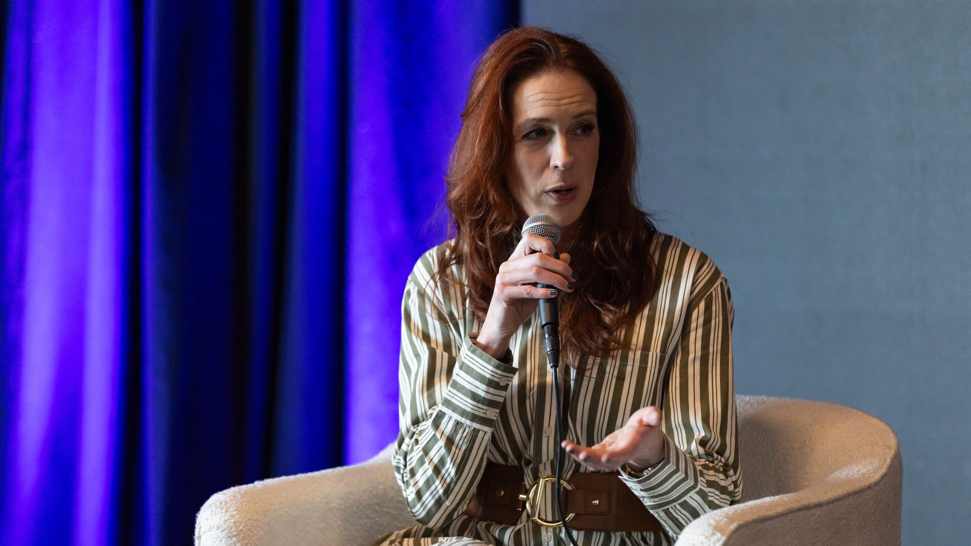 A woman with long red hair sits on a beige chair, holding a microphone, wearing a striped beige and white dress with a brown belt, speaking on a stage with purple curtains in the background.