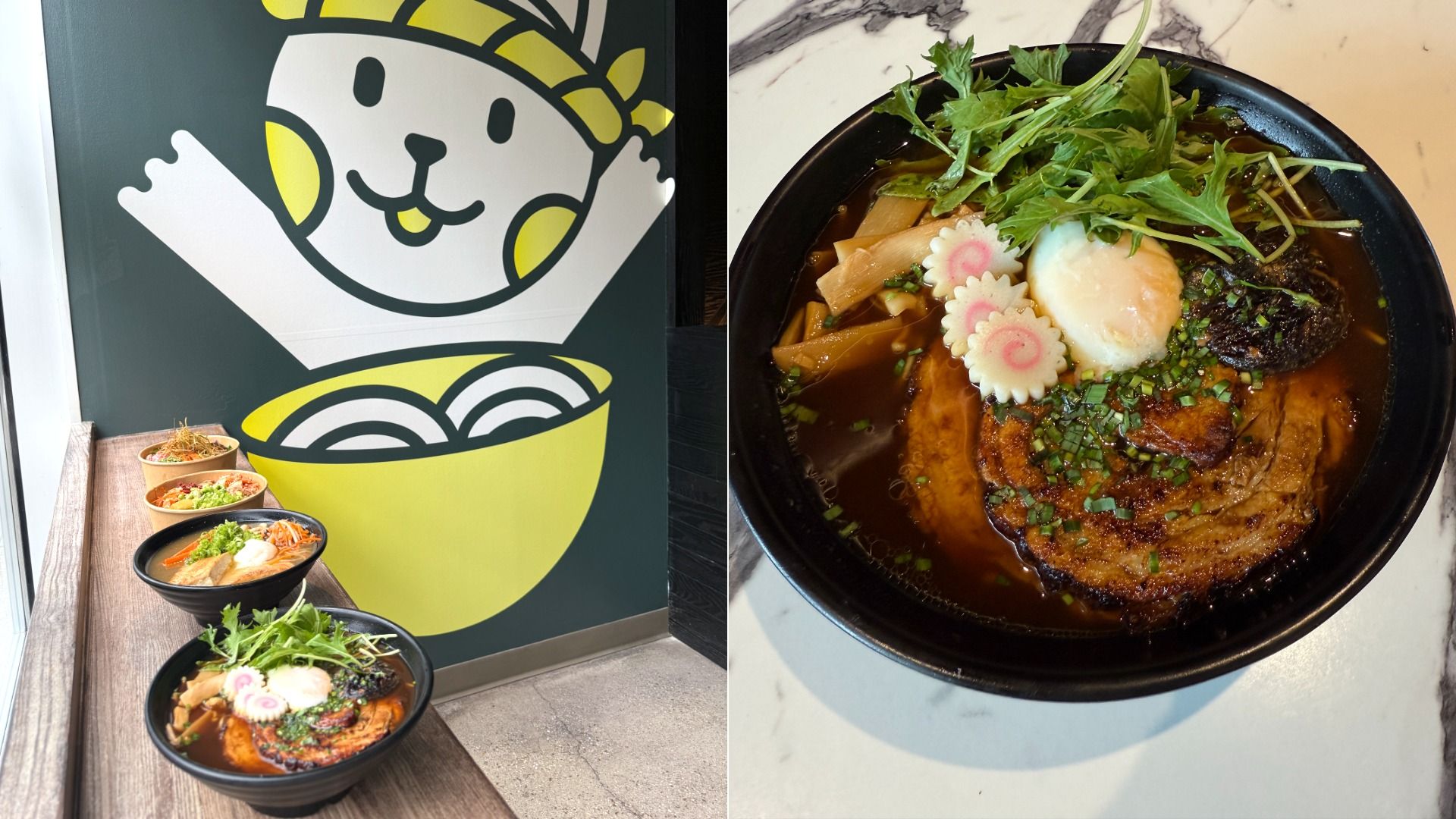 Two black bowls of ramen with green herbs, soft boiled eggs, fish cakes, and pork are placed on a wooden counter next to a wall with a cartoon cat in yellow and white holding a ramen bowl.