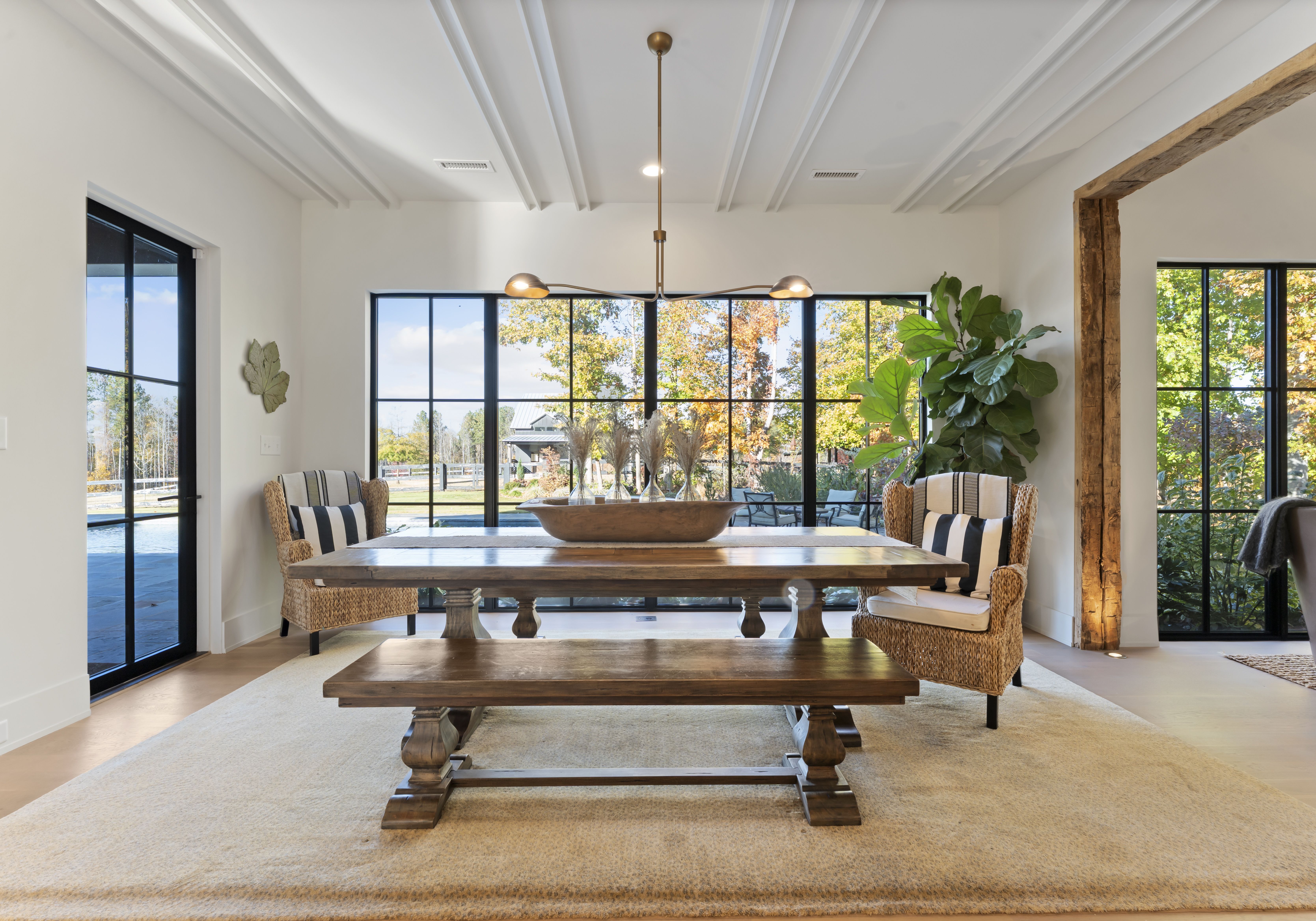 Bright dining room with a large wooden table and bench, wicker chairs with striped cushions, a branch chandelier, and tall black-framed windows showing autumn trees; a plant by a wooden beam.