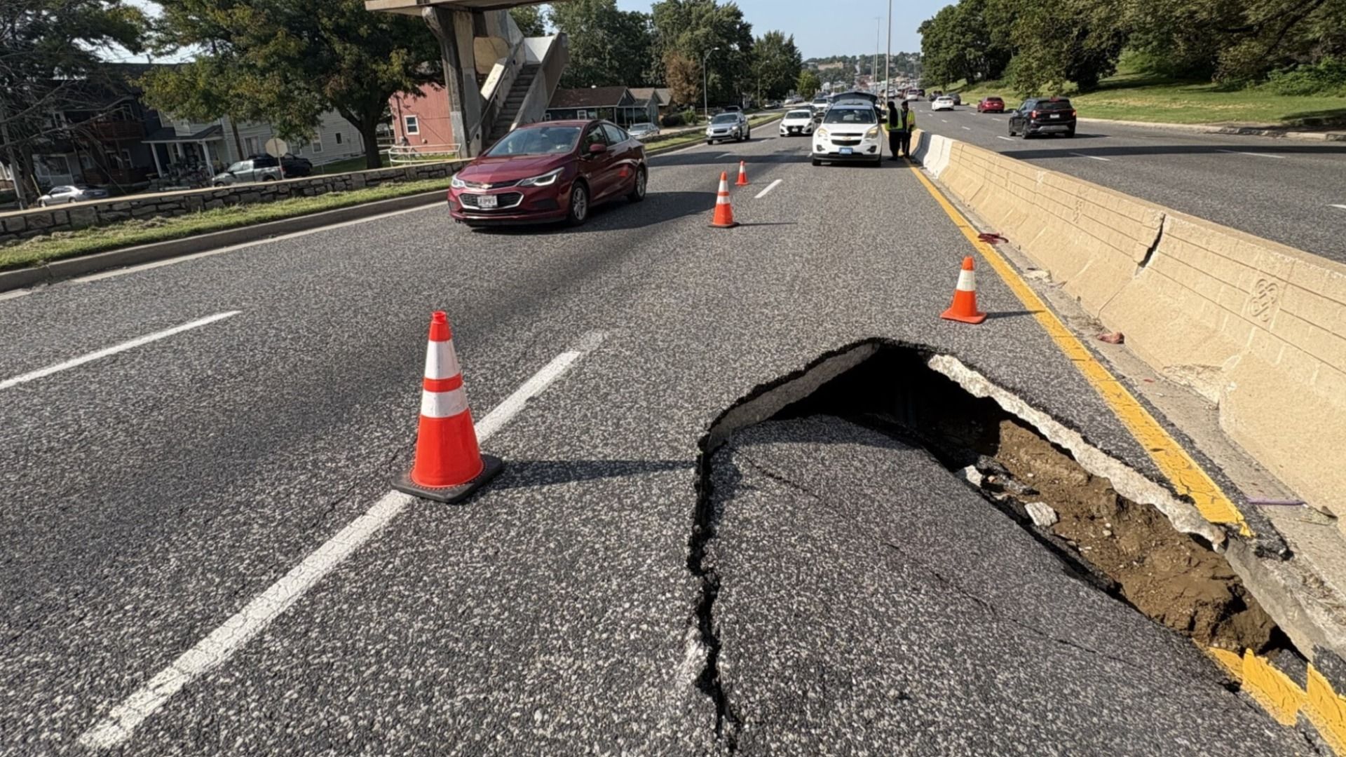 A multi-lane street with a large sinkhole in the right lane, blocked by orange traffic cones and a concrete barrier. Cars stop as two workers in reflective vests stand near a white car; trees and houses in the background.