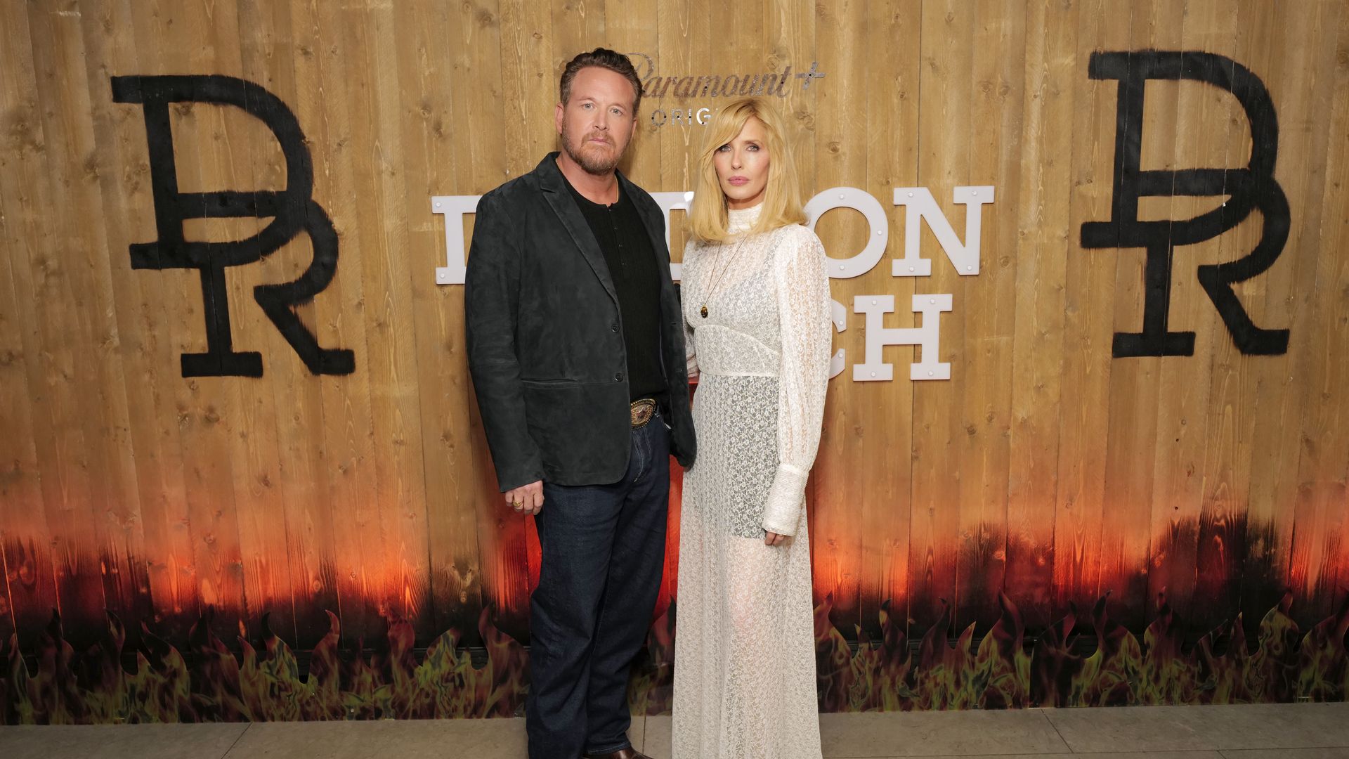Actors Cole Hauser and Kelly Reilly stand and post with serious faces in front of a "DUTTON RANCH" sign on wood.